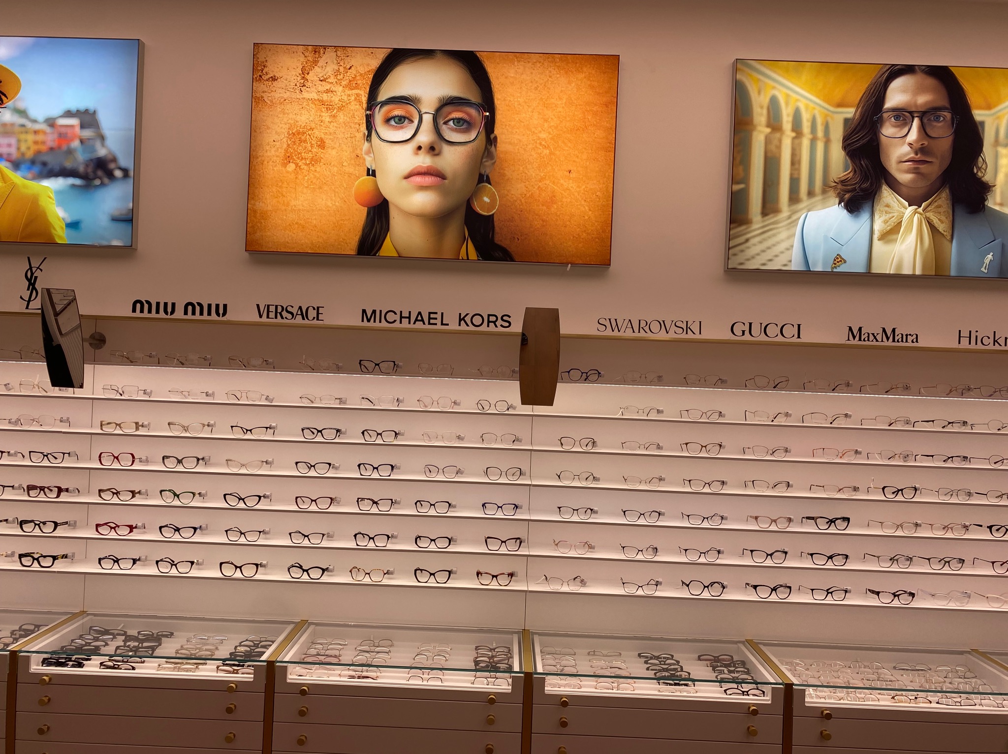 Salon optyczny: ekspozycja okularów różnych marek (Miu Miu, Versace, Michael Kors, Swarovski, Gucci, MaxMara) na białych półkach, z reklamami wizualnymi powyżej.