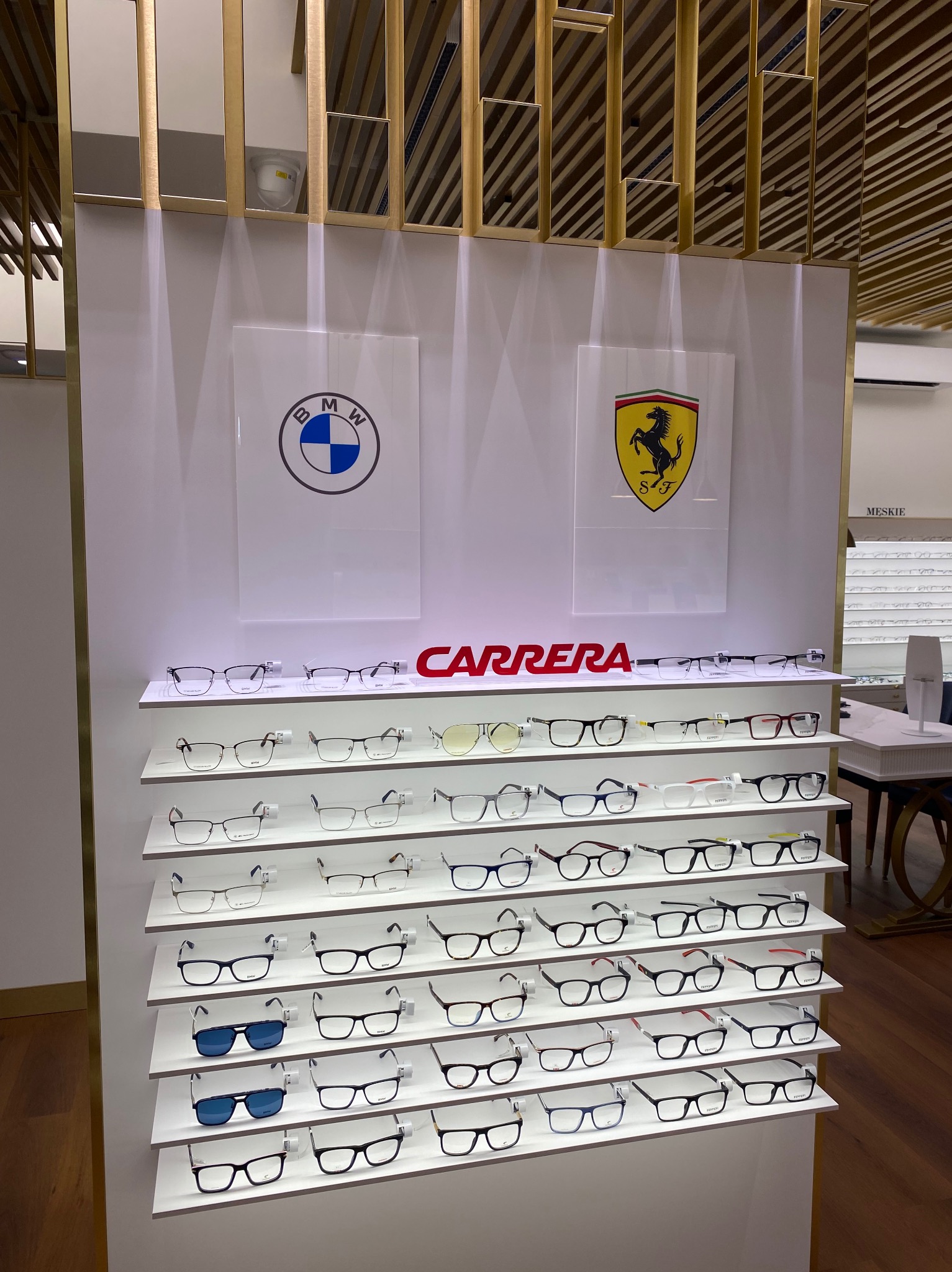 Ekspozycja okularów Carrera w salonie optycznym, z logo BMW i Ferrari w tle. Różne modele oprawek na białych półkach, oświetlenie punktowe, drewniana podłoga.