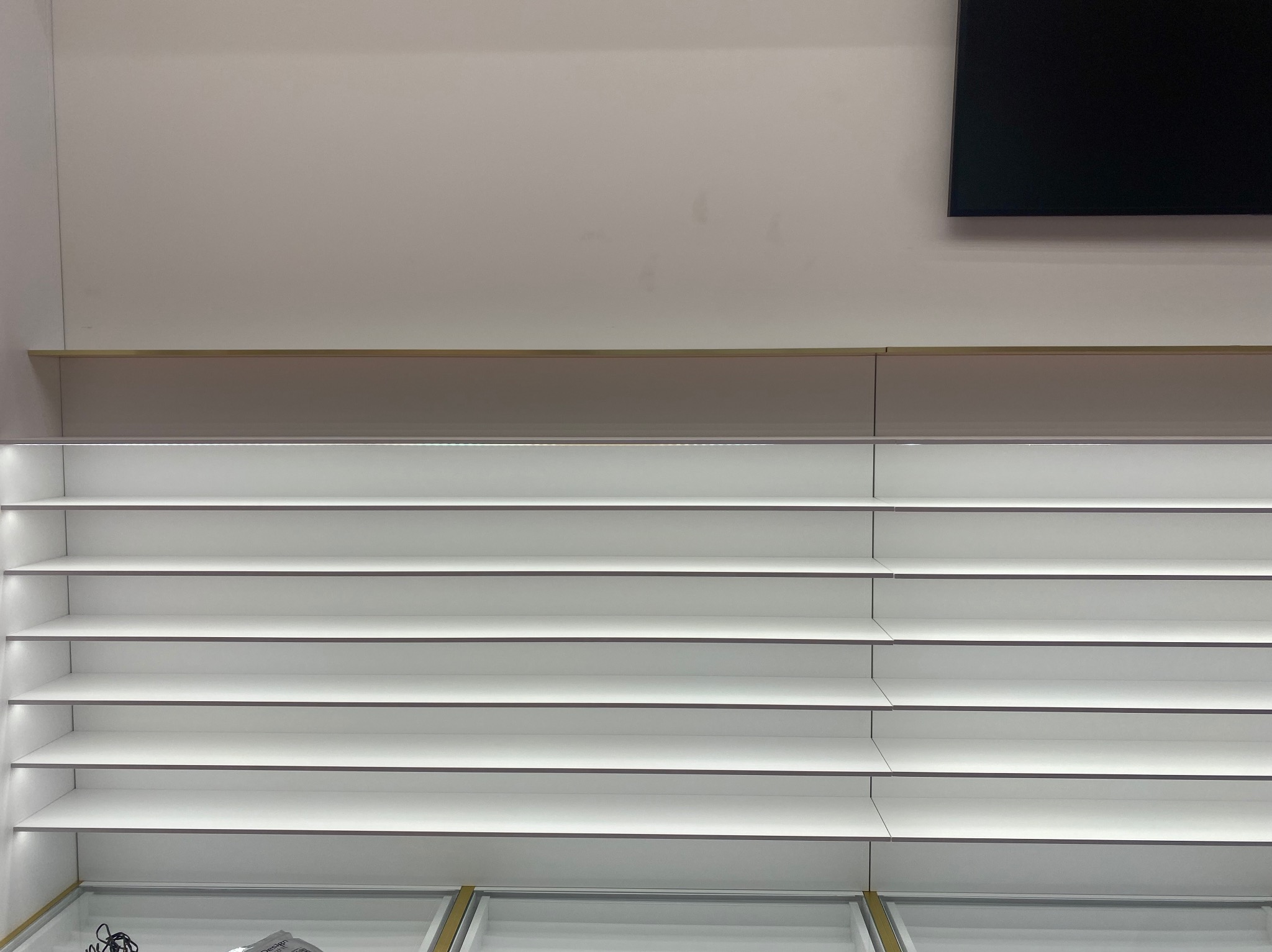 Białe, puste półki sklepowe z subtelnym oświetleniem LED, umieszczone pod ścianą z czarnym ekranem w górnym rogu. Minimalistyczny design.