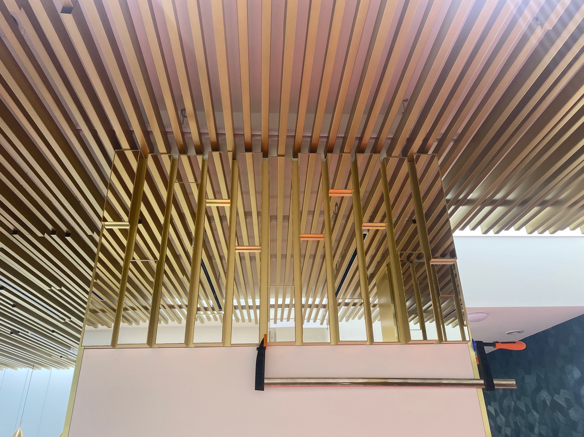 Złote, równoległe listwy sufitowe z lustrzanymi wstawkami, częściowo zamontowane, zabezpieczone ściskami stolarskimi. Widok z dołu, oświetlenie górne.