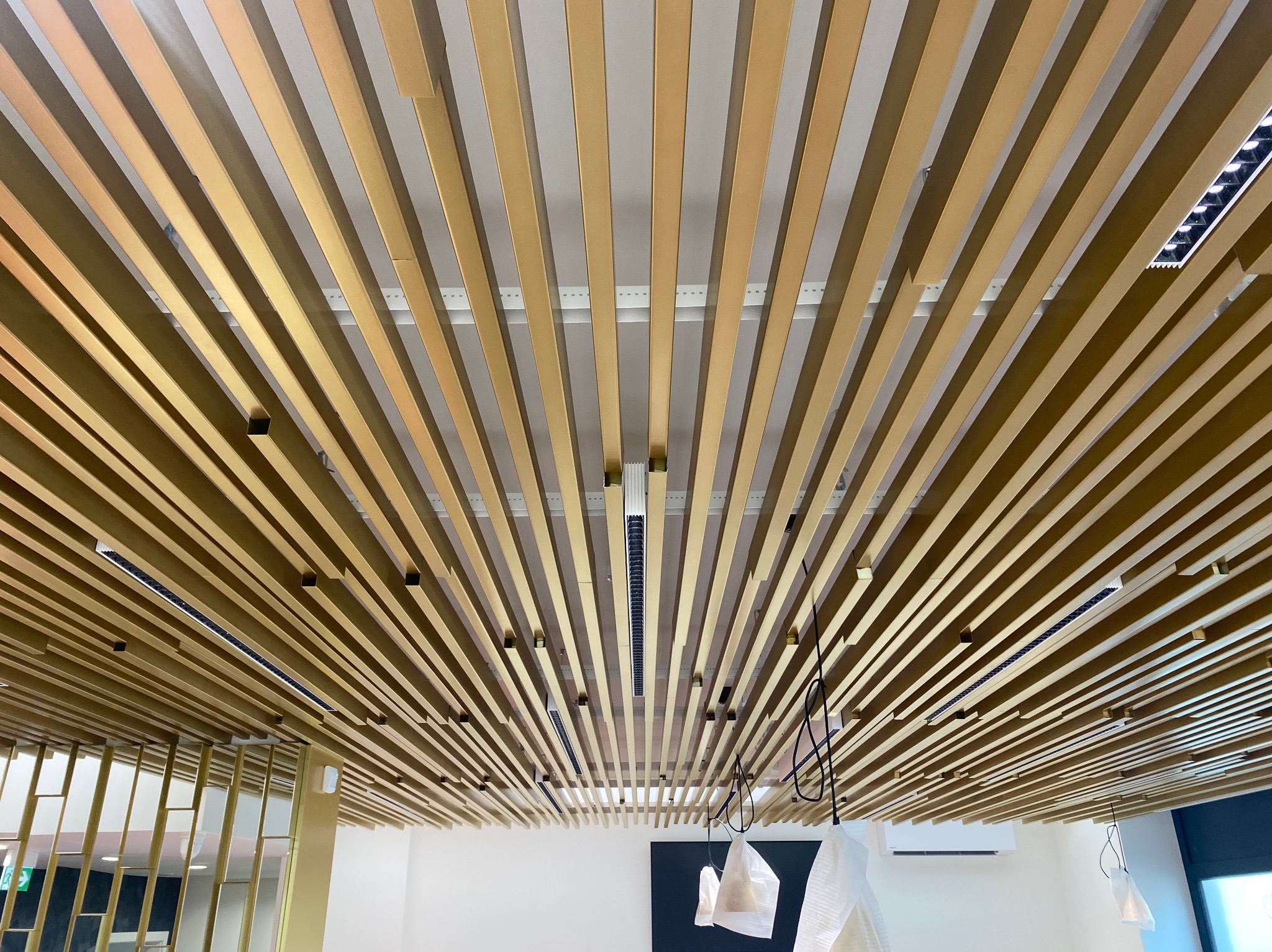 Dekoracyjny sufit z drewnianymi listwami i wbudowanym oświetleniem LED, widok z dołu. Częściowo widoczne elementy instalacji i klimatyzacji.