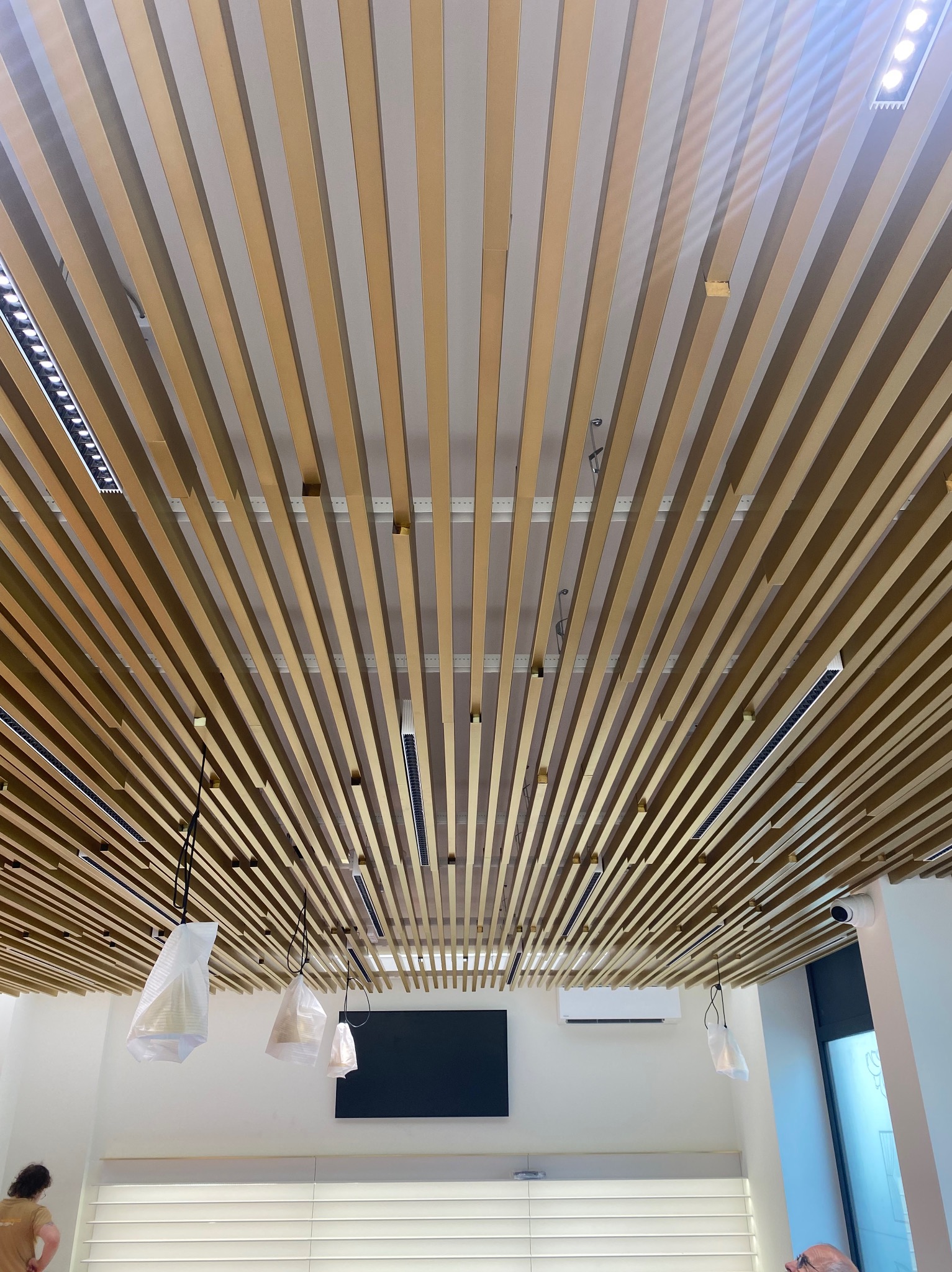 Dekoracyjny sufit z drewnianych listew i zamontowanym oświetleniem LED. Lampy wiszące w folii ochronnej. Widoczny klimatyzator i kamera przemysłowa.