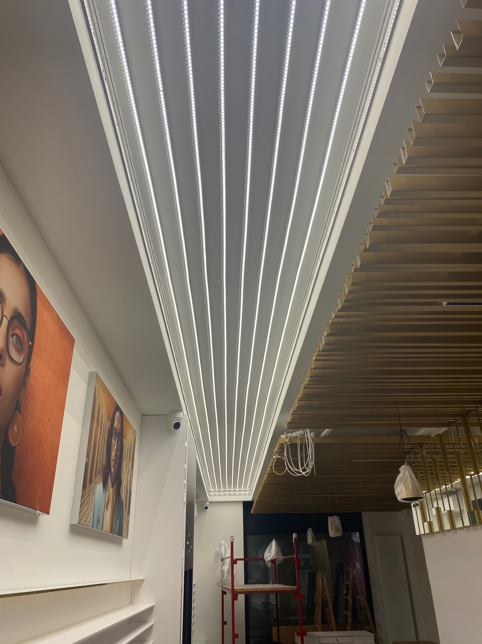 Nowoczesne oświetlenie sufitowe LED z równoległymi pasami światła w jasnym wnętrzu sklepu, widoczne elementy dekoracyjne i fragmenty plakatów na ścianach.