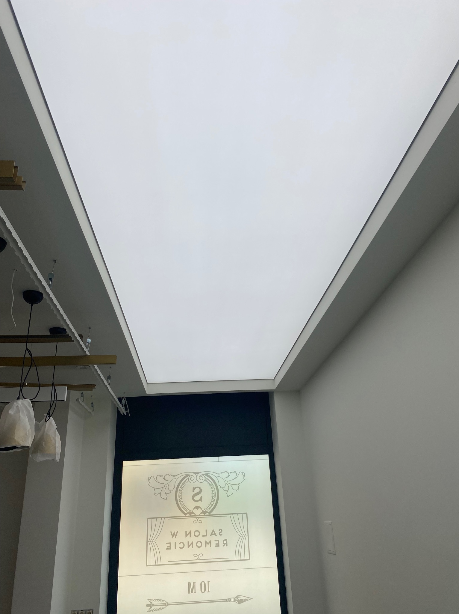 Biały sufit napinany z oświetleniem LED w salonie, widoczne lampy wiszące i dekoracyjne elementy wykończenia wnętrza. Minimalistyczny design.