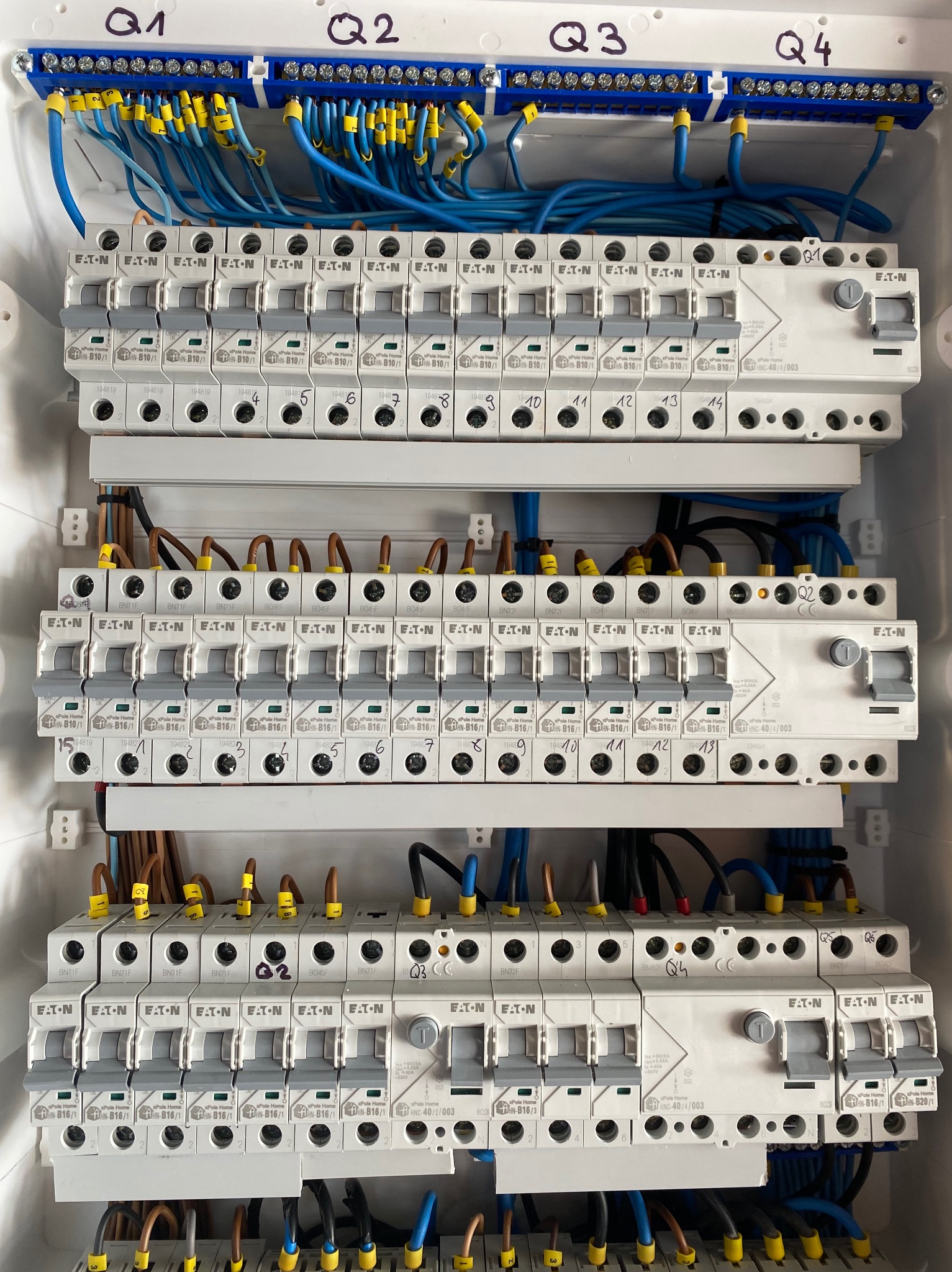 Rozdzielnica elektryczna z zamontowanymi bezpiecznikami Eaton, oznaczonymi numerami i podłączonymi przewodami w kolorach niebieskim, brązowym i czarnym. Widoczne oznaczenia Q1-Q4.