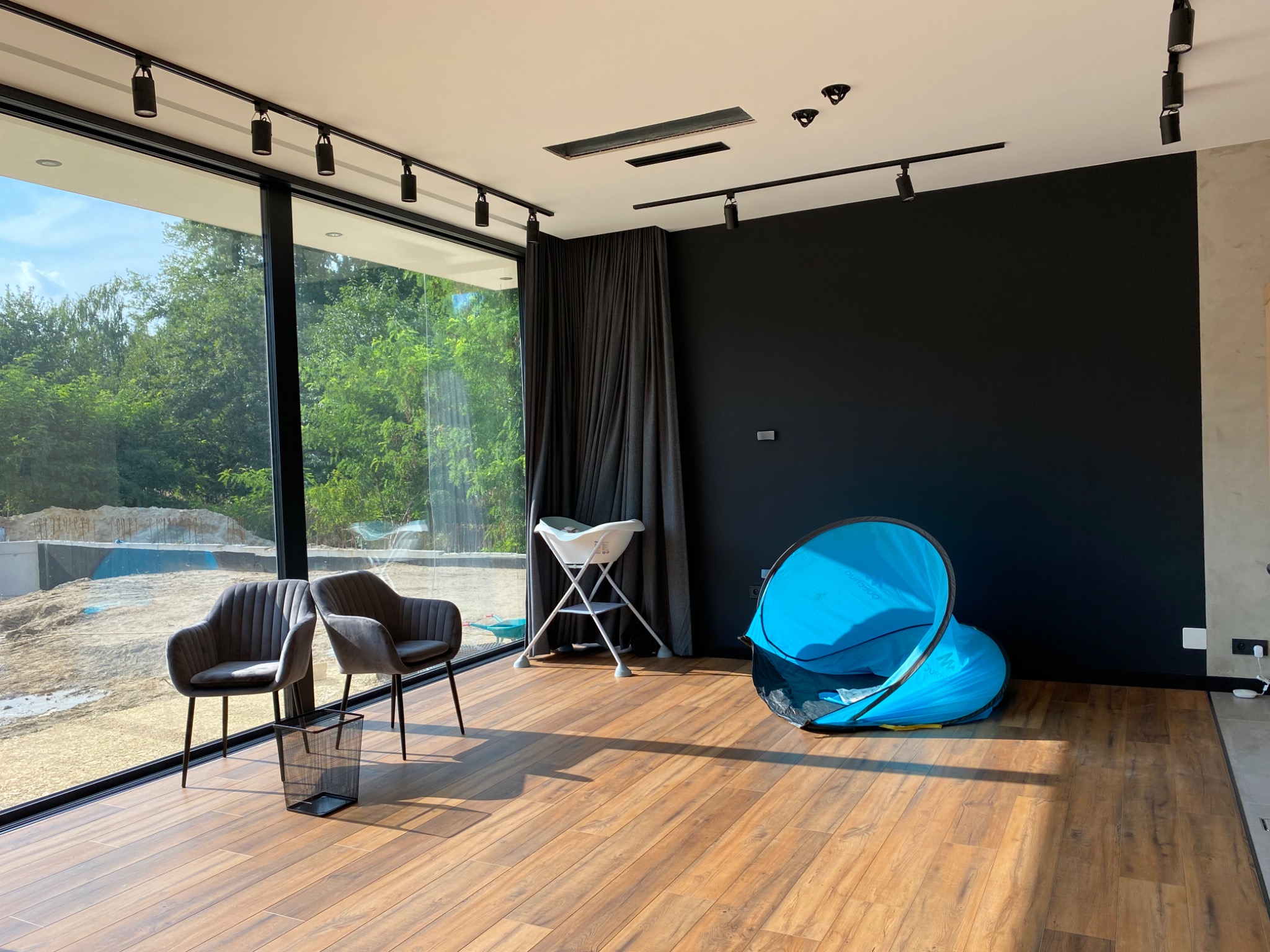 Nowoczesne wnętrze z drewnianą podłogą i oknem panoramicznym, oświetlone reflektorami na szynoprzewodach. Widoczne dwa fotele i elementy wyposażenia.