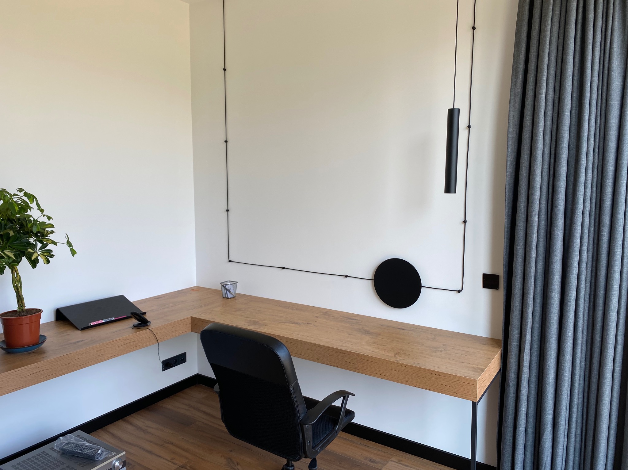 Nowoczesne biuro domowe z drewnianym biurkiem, czarnym krzesłem i minimalistycznym oświetleniem punktowym na ścianie. Roślina doniczkowa dodaje naturalnego akcentu.