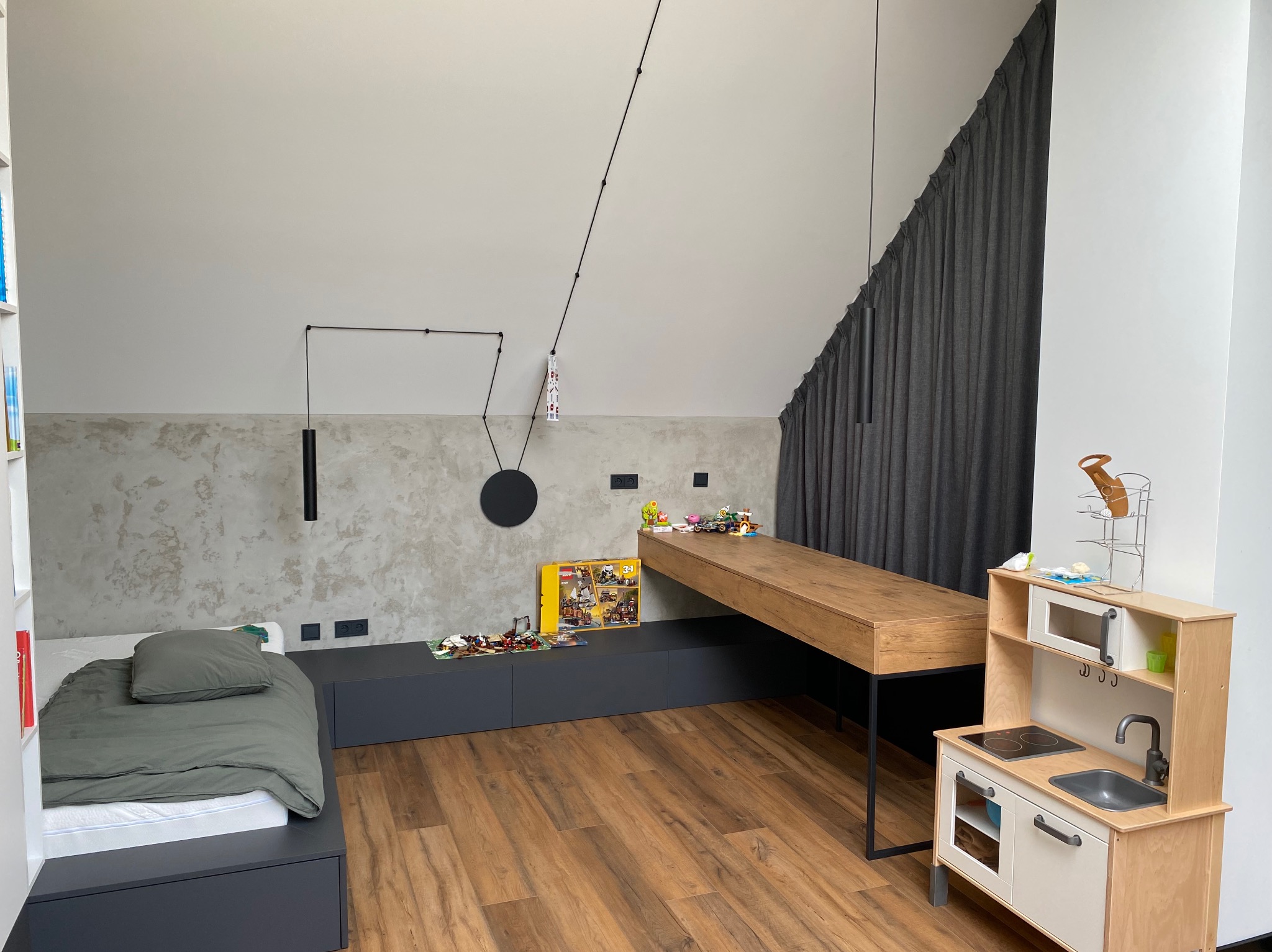 Pokój dziecięcy na poddaszu z drewnianą podłogą, szarą zabudową, biurkiem i lampą wiszącą. Widoczne gniazdka elektryczne na ścianie.