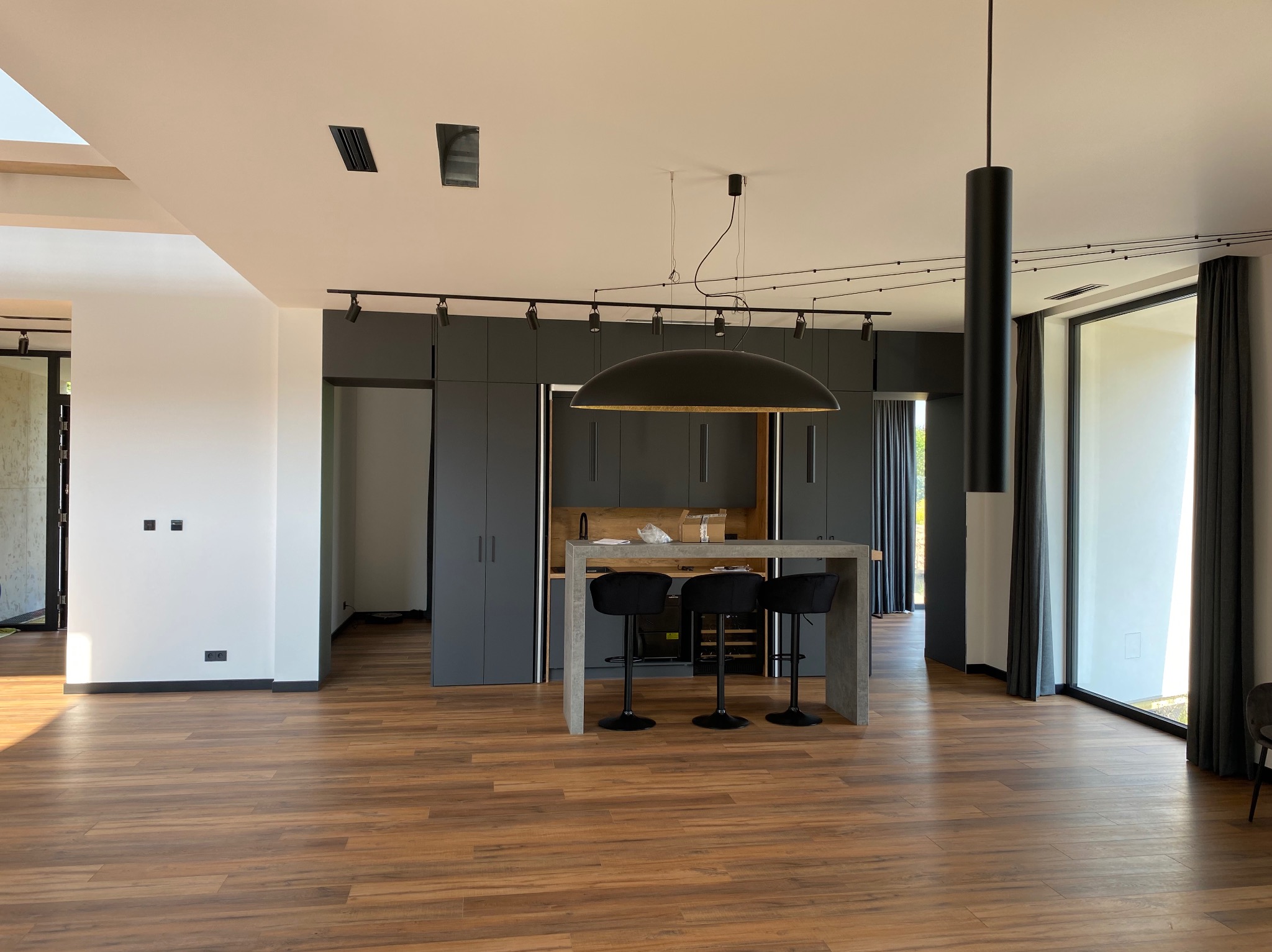 Nowoczesne wnętrze z drewnianą podłogą, szarą zabudową kuchenną i wyspą. Widoczne oświetlenie szynowe i wisząca lampa nad wyspą. Minimalistyczny design.