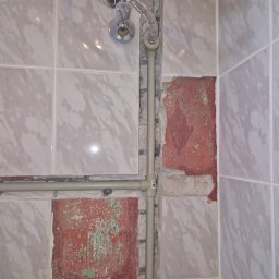 Gheorghe Gavriciuc - Fragment ściany z częściowo usuniętymi płytkami, odsłaniający starą czerwoną cegłę i fragment instalacji wodnej z rurami. Widoczne białe płytki imitujące marmur.