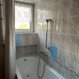 Gheorghe Gavriciuc - Wykończona łazienka z wanną, szarymi płytkami imitującymi marmur i niebieskimi akcentami. Widoczna bateria wannowa, okno z firanką i suszarka podsufitowa.