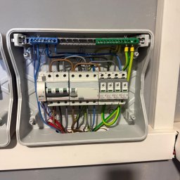 Gheorghe Gavriciuc - Szczegółowe ujęcie wnętrza skrzynki elektrycznej z zainstalowanymi bezpiecznikami i przewodami o różnych kolorach, zamontowanej na szarej ścianie.