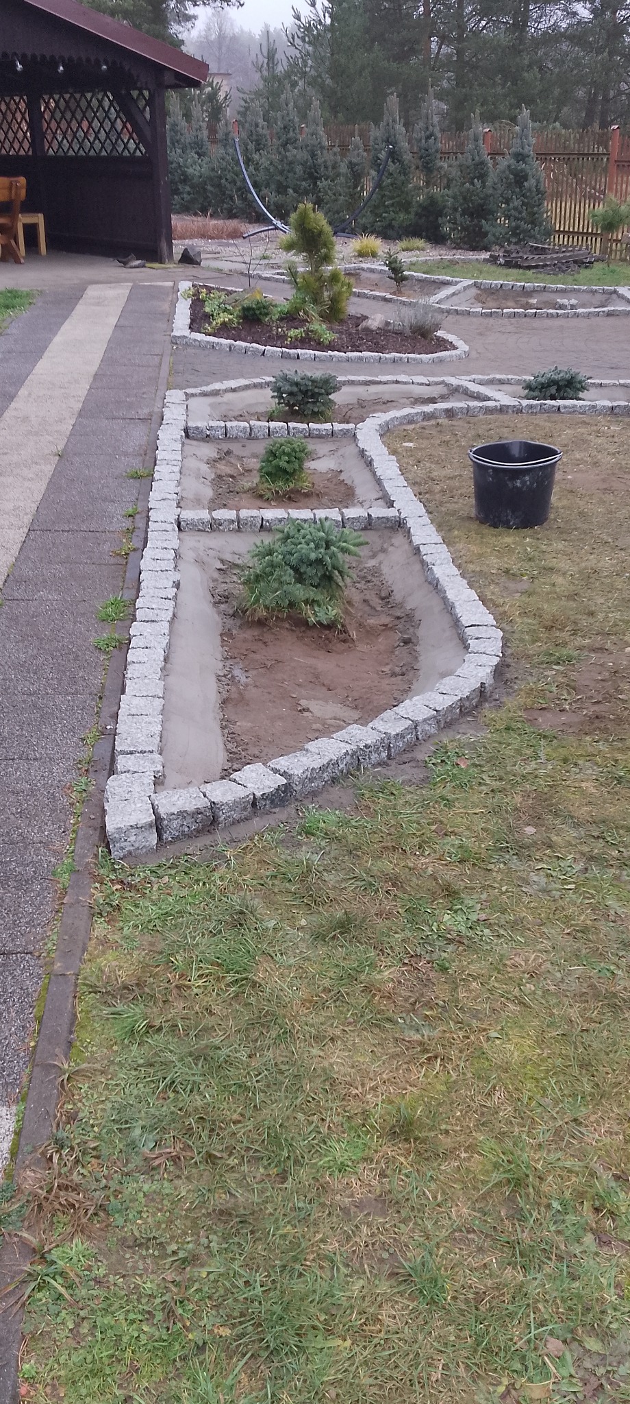 Kaskadowe rabaty z obrzeżami z kostki granitowej, świeżo po ułożeniu, z młodymi iglakami. Widoczny fragment chodnika i altana w tle. Prace brukarskie w ogrodzie.