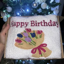 Aleksandra Hinc - Biały ręcznik z haftowanym bukietem tulipanów i napisem 'Happy Birthday' w pudełku, na tle choinki z lampkami. Prezent urodzinowy z unikalnym haftem.