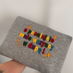 Aleksandra Hinc - Szara bluza z kolorowym haftem napisu 'a pocałujcie mnie wszyscy w dupę!' i motywem gwiazdek, trzymana w dłoni, na jasnym tle.