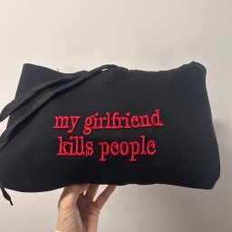 Aleksandra Hinc - Czarna bluza z kapturem, trzymana w dłoni, z czerwonym haftem 'my girlfriend kills people'. Widoczne sznurki kaptura i fragment ręki.