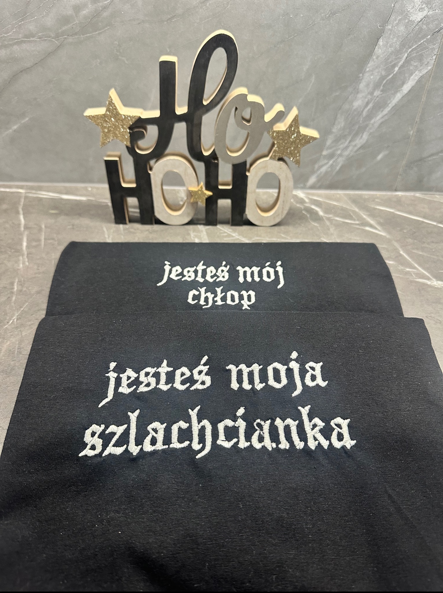 Dwa czarne T-shirty z białym haftem: 'jesteś mój chłop' i 'jesteś moja szlachcianka'. W tle dekoracja 'Ho Ho Ho' z gwiazdkami.