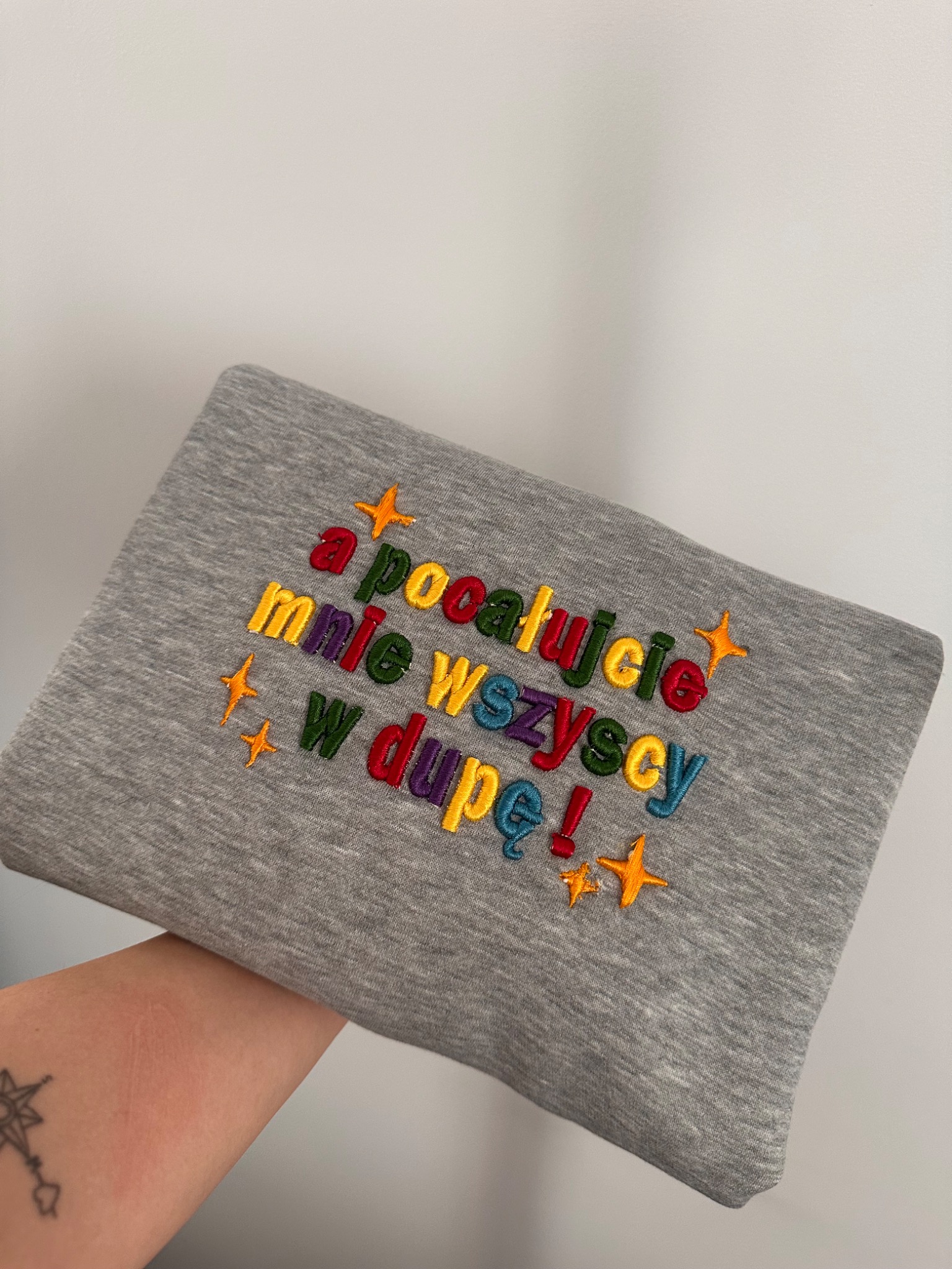 Szara bluza z kolorowym haftem napisu 'a pocałujcie mnie wszyscy w dupę!' i motywem gwiazdek, trzymana w dłoni, na jasnym tle.