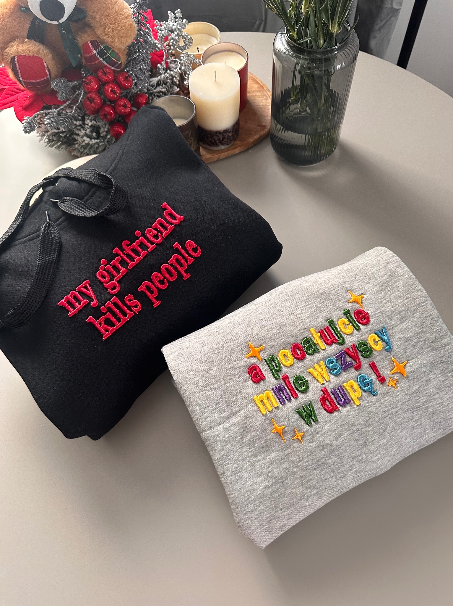 Dwie złożone bluzy z kapturem: czarna z czerwonym haftem 'my girlfriend kills people' i szara z kolorowym haftem w języku polskim, leżą na stole.