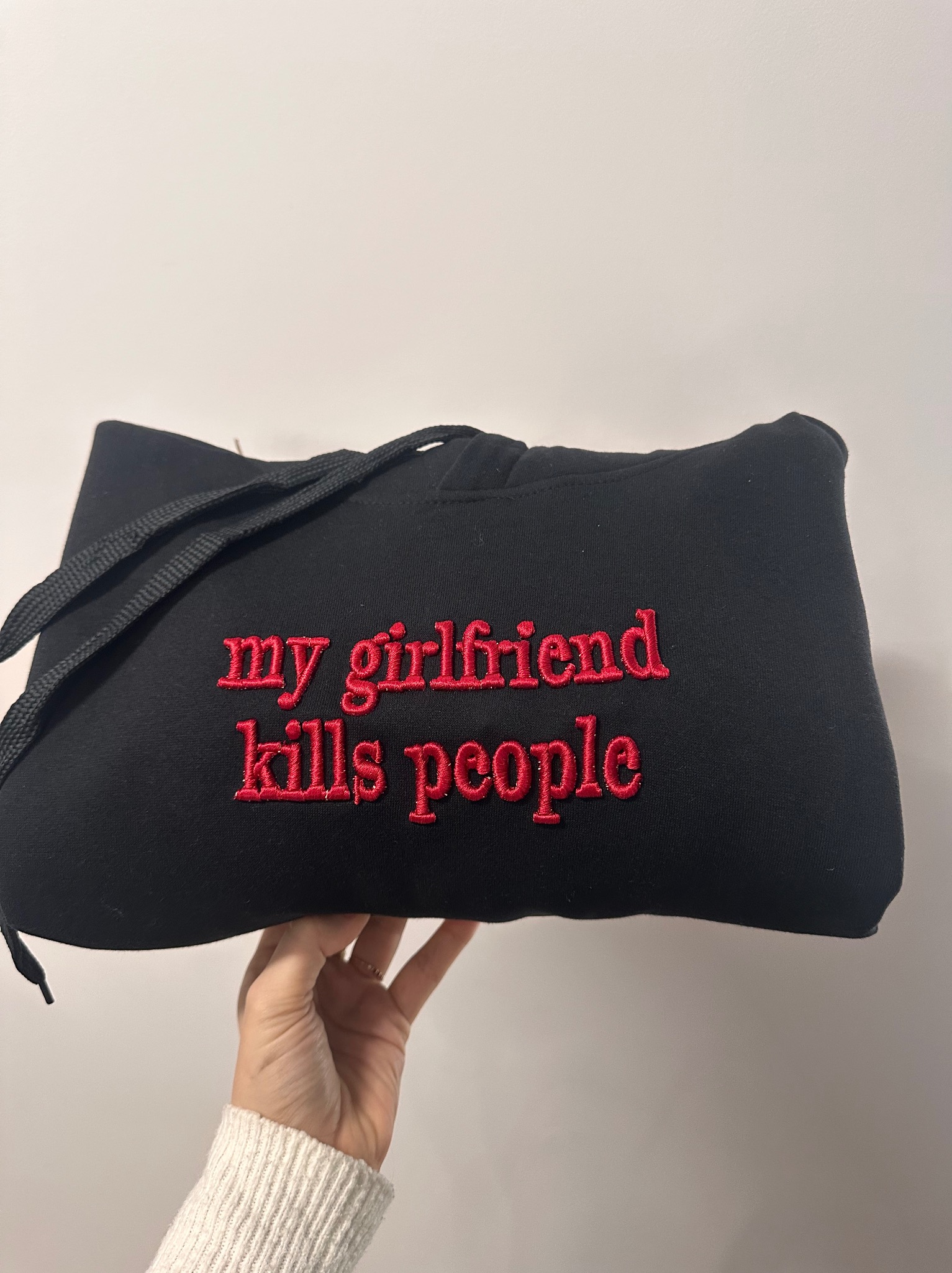 Czarna bluza z kapturem, trzymana w dłoni, z czerwonym haftem 'my girlfriend kills people'. Widoczne sznurki kaptura i fragment ręki.