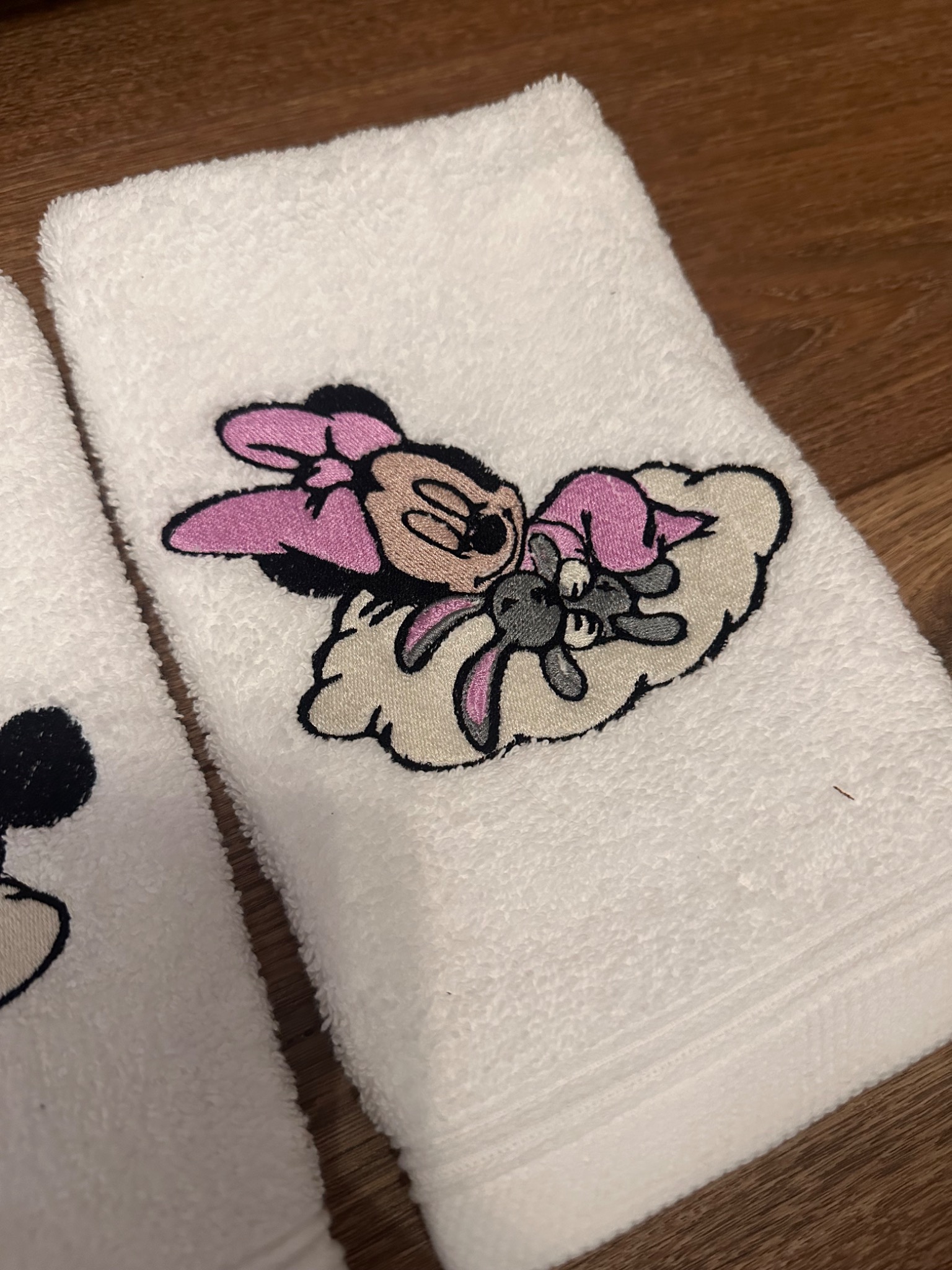 Biały ręcznik frotte z haftem postaci z bajek, leżący na drewnianej powierzchni. Widoczne detale haftu i faktura materiału.