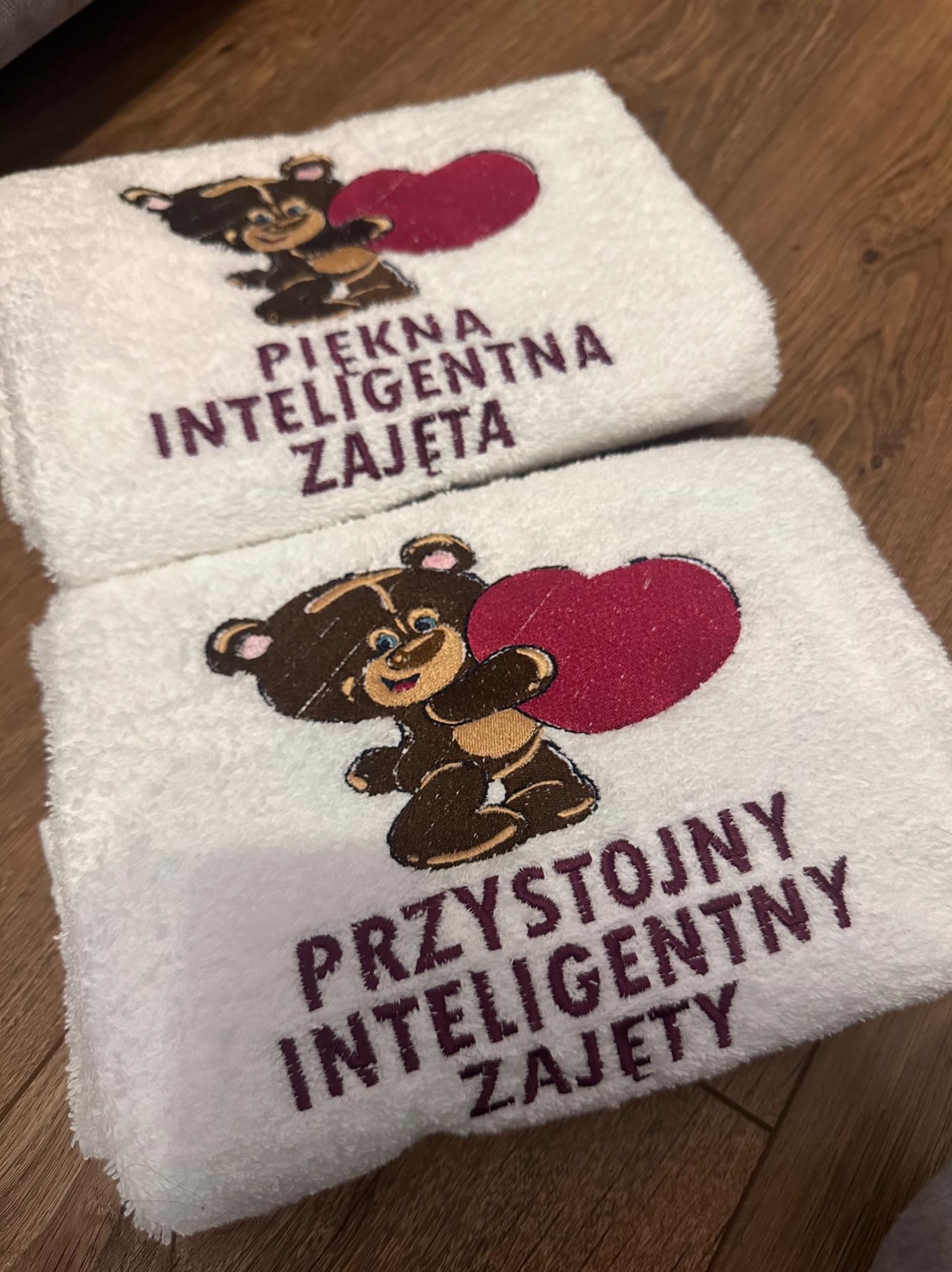Dwa białe ręczniki z haftem: jeden z misiem i napisem 'Piękna Inteligentna Zajęta', drugi z misiem i napisem 'Przystojny Inteligentny Zajęty'.