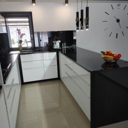 Adrian Hofman - Nowoczesna kuchnia z białymi szafkami i czarnym blatem. Na blacie misa z owocami, w tle okno z kwiatami i designerski zegar na ścianie. Minimalistyczny styl.