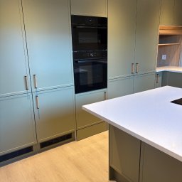 Adrian Hofman - Nowoczesna kuchnia w odcieniach zieleni z zabudową meblową pod sufit, wbudowanym piekarnikiem i wyspą z białym blatem. Widoczna drewniana podłoga i minimalistyczne uchwyty.