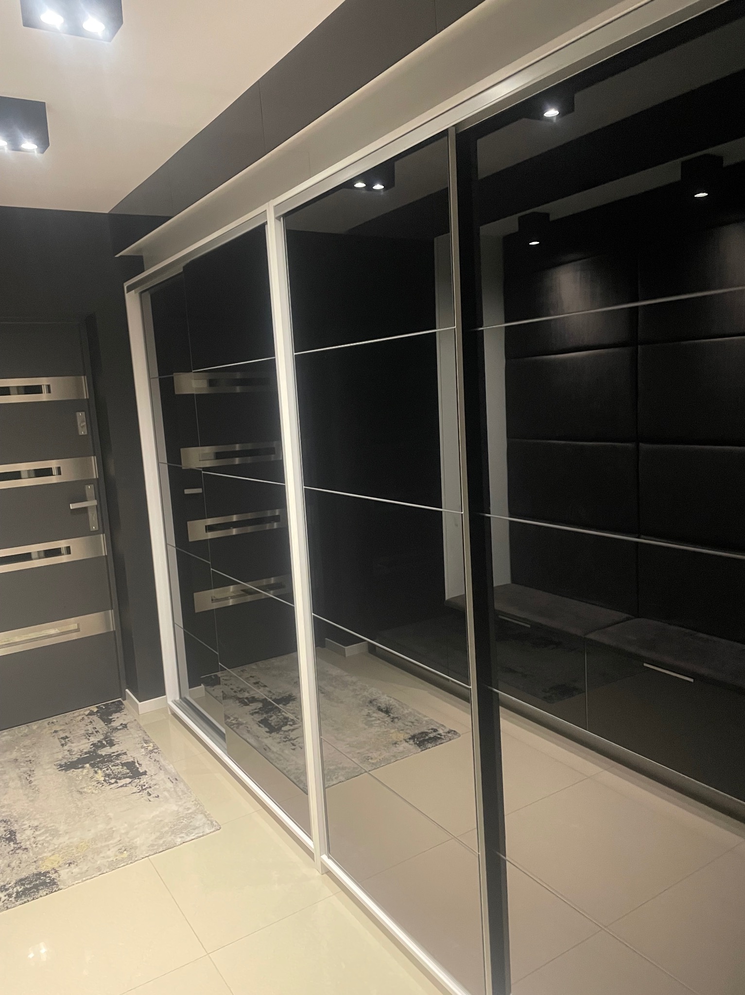 Nowoczesna szafa przesuwna z lustrzanymi drzwiami w ciemnym wnętrzu. Widoczny fragment dywanu i drzwi wejściowe. Minimalistyczny design, eleganckie wykończenie.