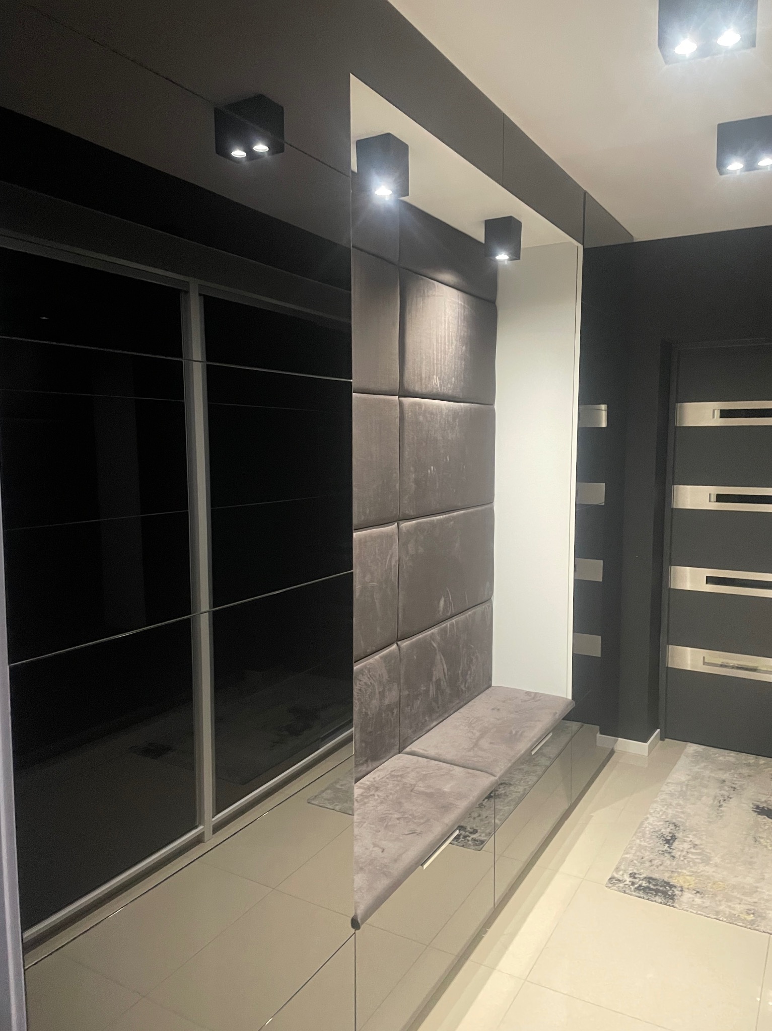 Nowoczesny przedpokój w odcieniach szarości i bieli. Szafa przesuwna z lustrzanymi drzwiami, tapicerowany panel ścienny z siedziskiem i punktowe oświetlenie sufitowe.