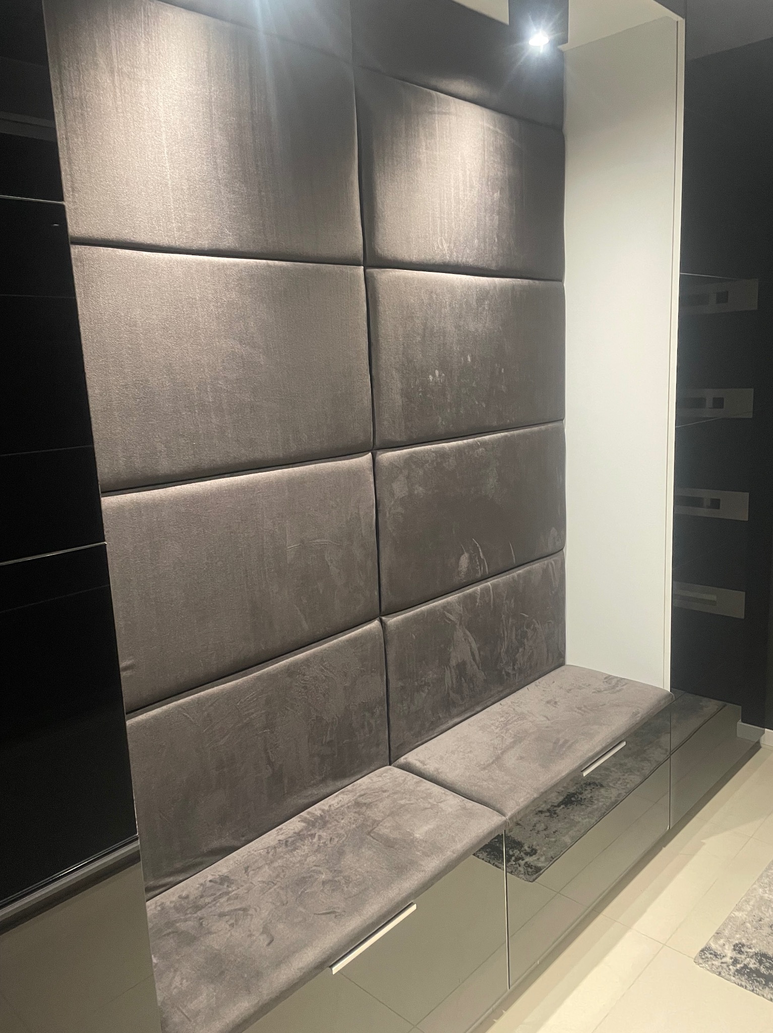 Tapicerowany panel ścienny w odcieniach szarości z siedziskiem, tworzący nowoczesny i elegancki element wystroju wnętrza. Widoczne detale wykończenia i oświetlenie.