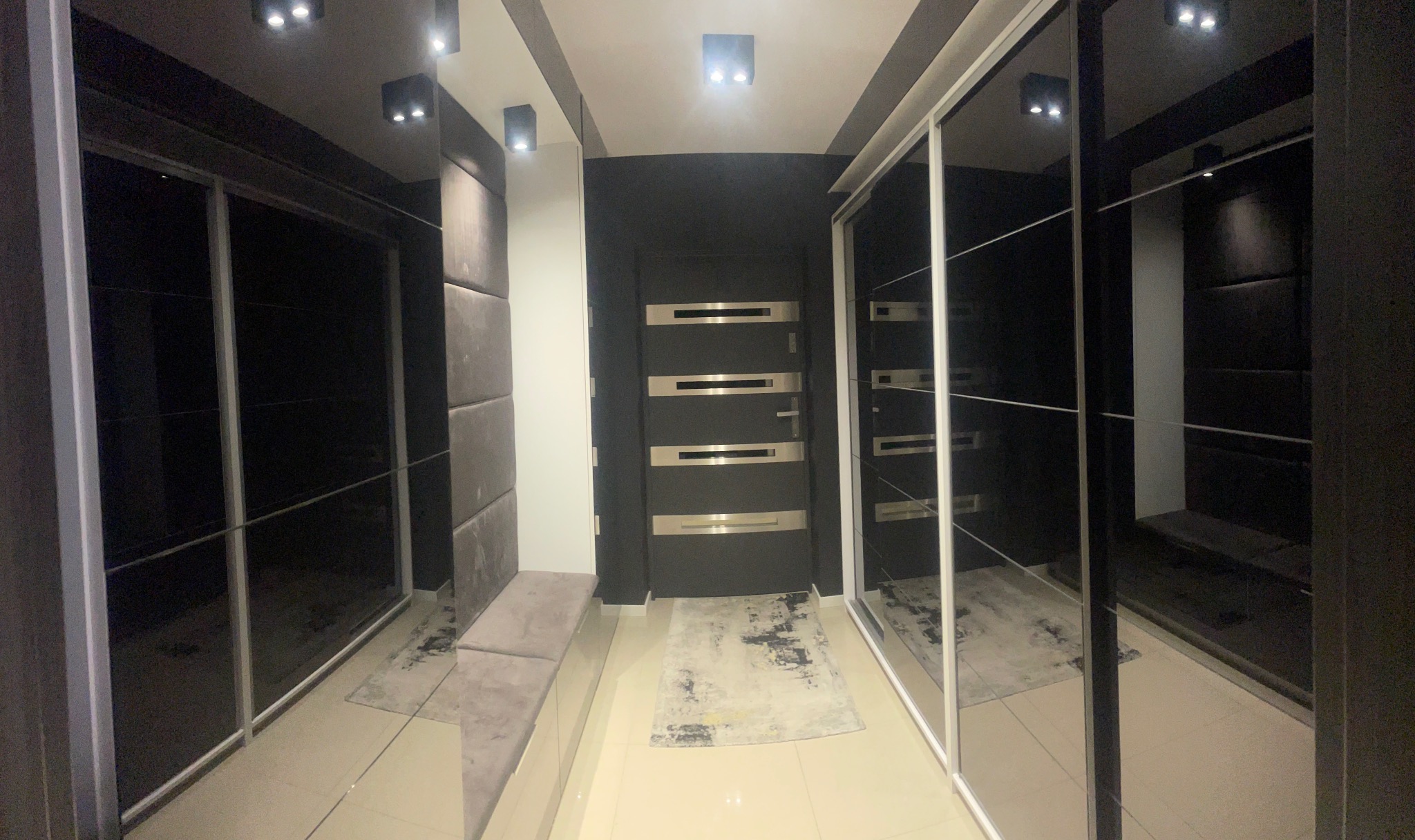 Wnętrze wiatrołapu z zabudową szafy przesuwnej z lustrami, tapicerowanym siedziskiem i drzwiami wejściowymi w ciemnej tonacji, oświetlone punktowo.