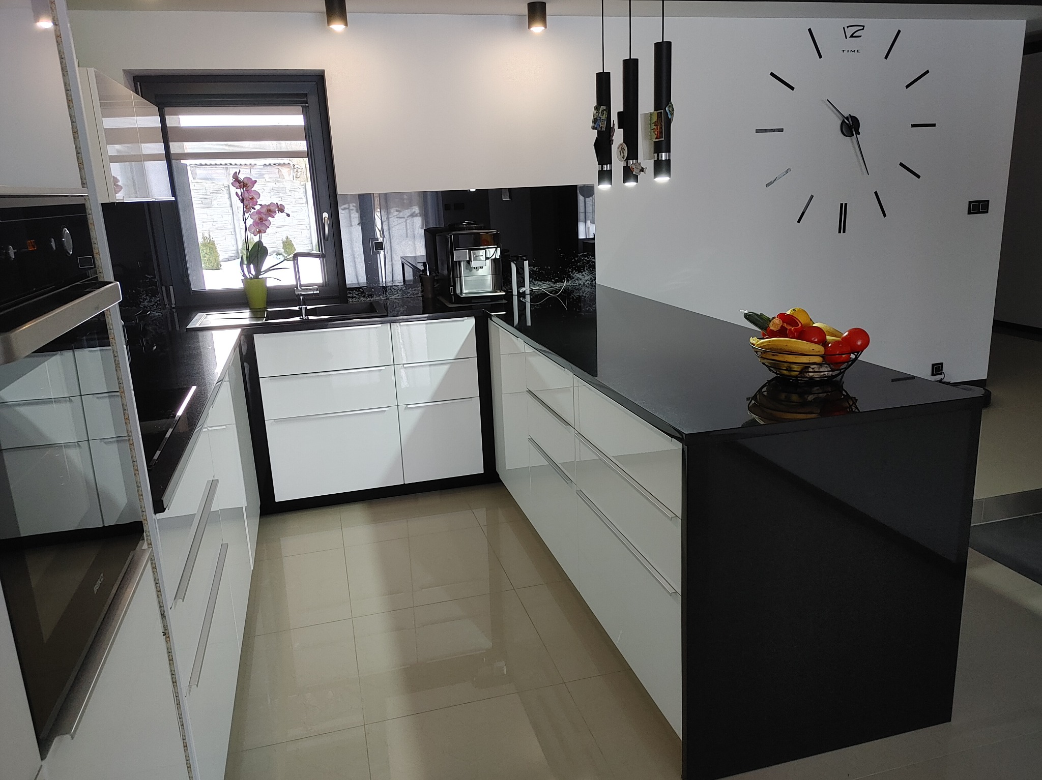 Nowoczesna kuchnia z białymi szafkami i czarnym blatem. Na blacie misa z owocami, w tle okno z kwiatami i designerski zegar na ścianie. Minimalistyczny styl.