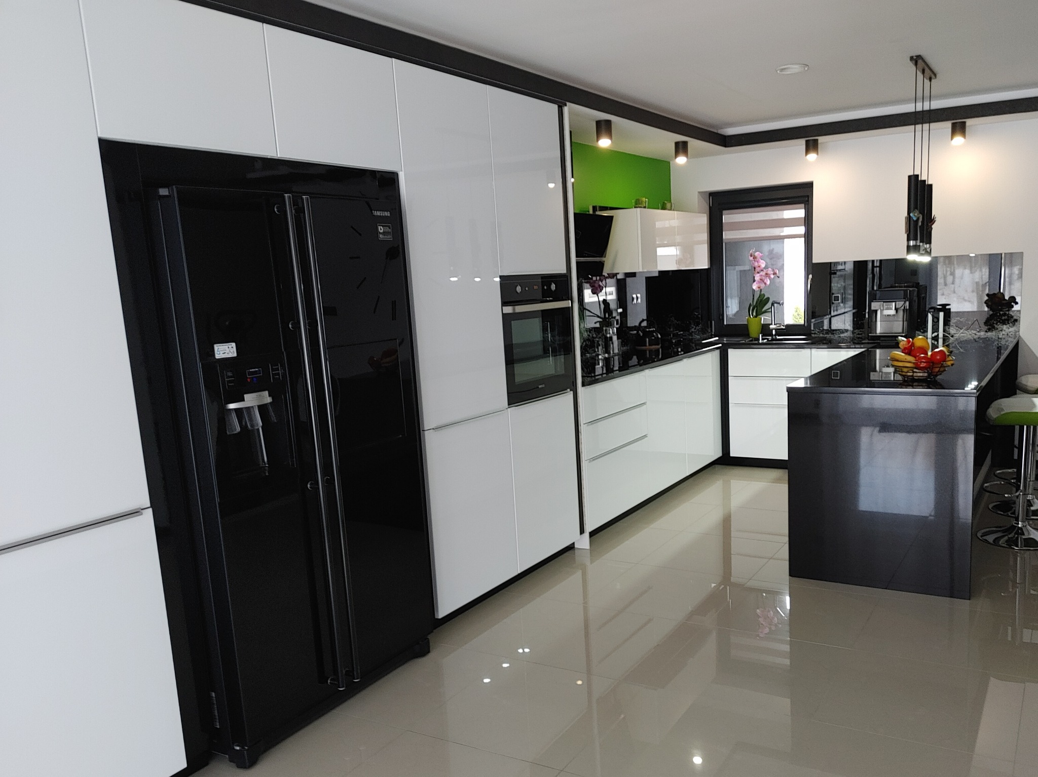 Nowoczesna kuchnia z białymi szafkami, czarnym blatem i lodówką Samsung z dystrybutorem wody. Zielona ściana w tle dodaje akcentu kolorystycznego.