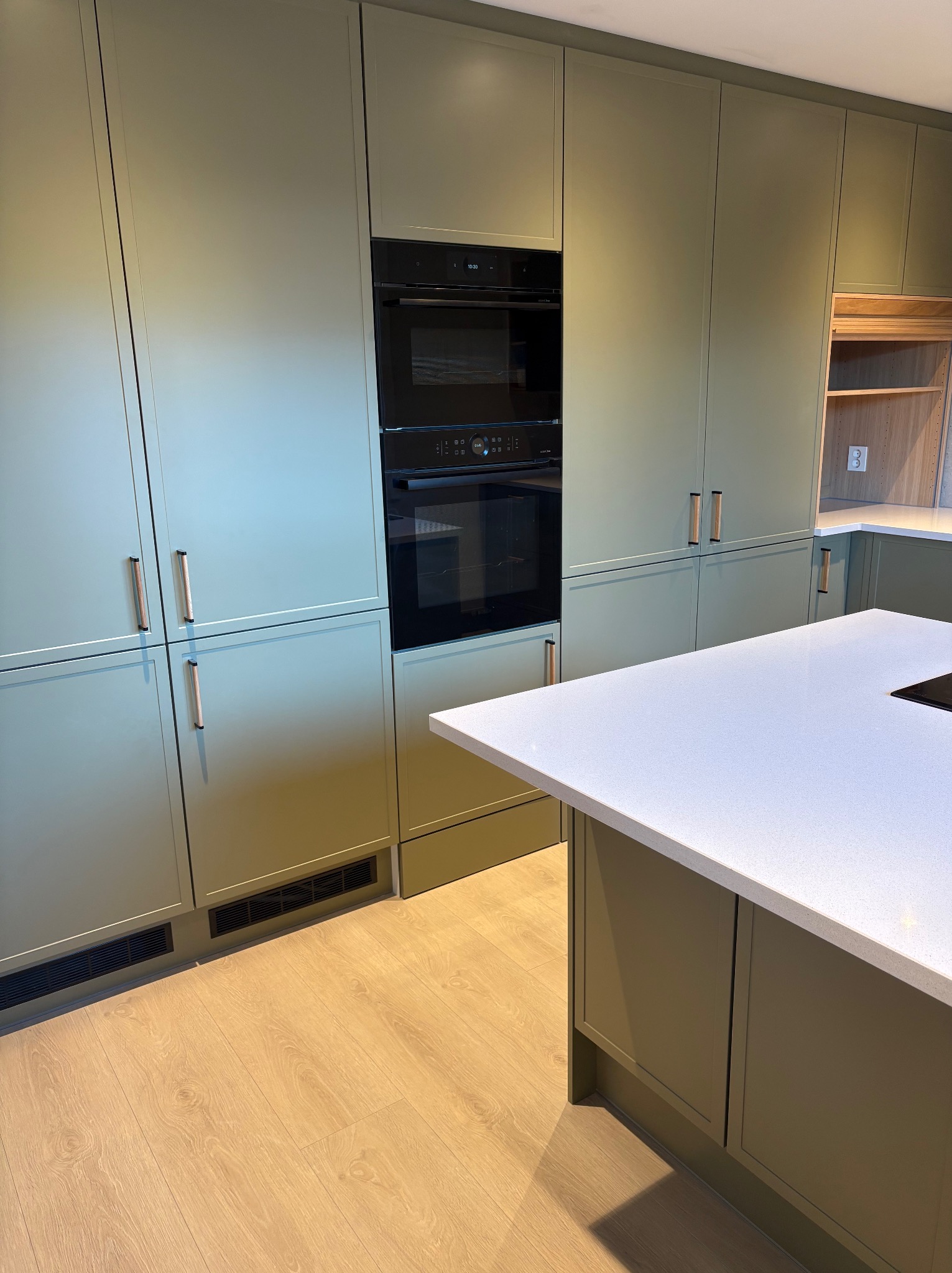 Nowoczesna kuchnia w odcieniach zieleni z zabudową meblową pod sufit, wbudowanym piekarnikiem i wyspą z białym blatem. Widoczna drewniana podłoga i minimalistyczne uchwyty.