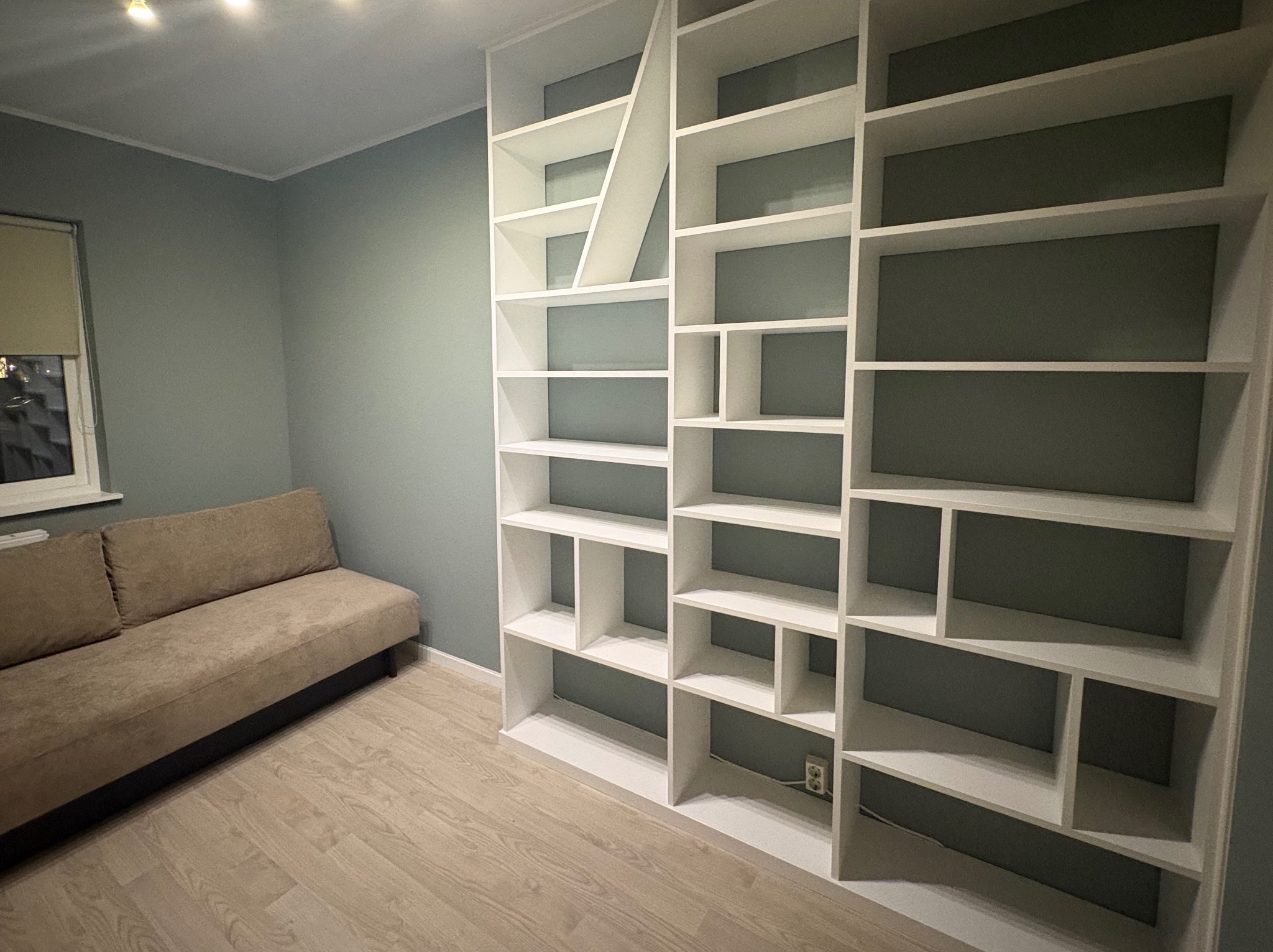 Biały, asymetryczny regał na książki z półkami o różnych kształtach, stojący w pokoju z szarozielonymi ścianami i beżową sofą. Widoczne gniazdko elektryczne przy regale.