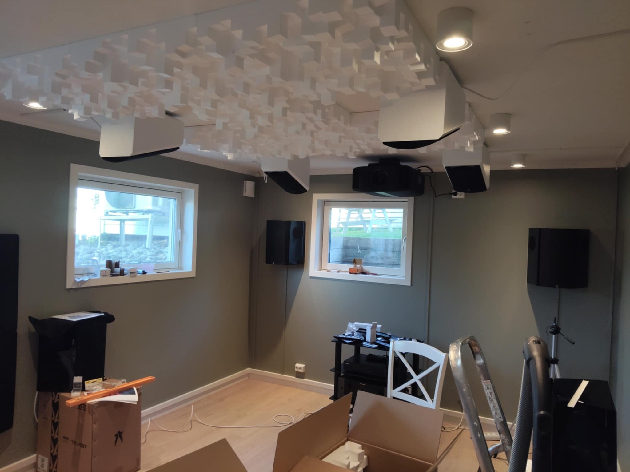 Wygłuszone pomieszczenie kina domowego z panelami akustycznymi na suficie, głośnikami i projektorem. Widoczne okna, drabina i kartony.