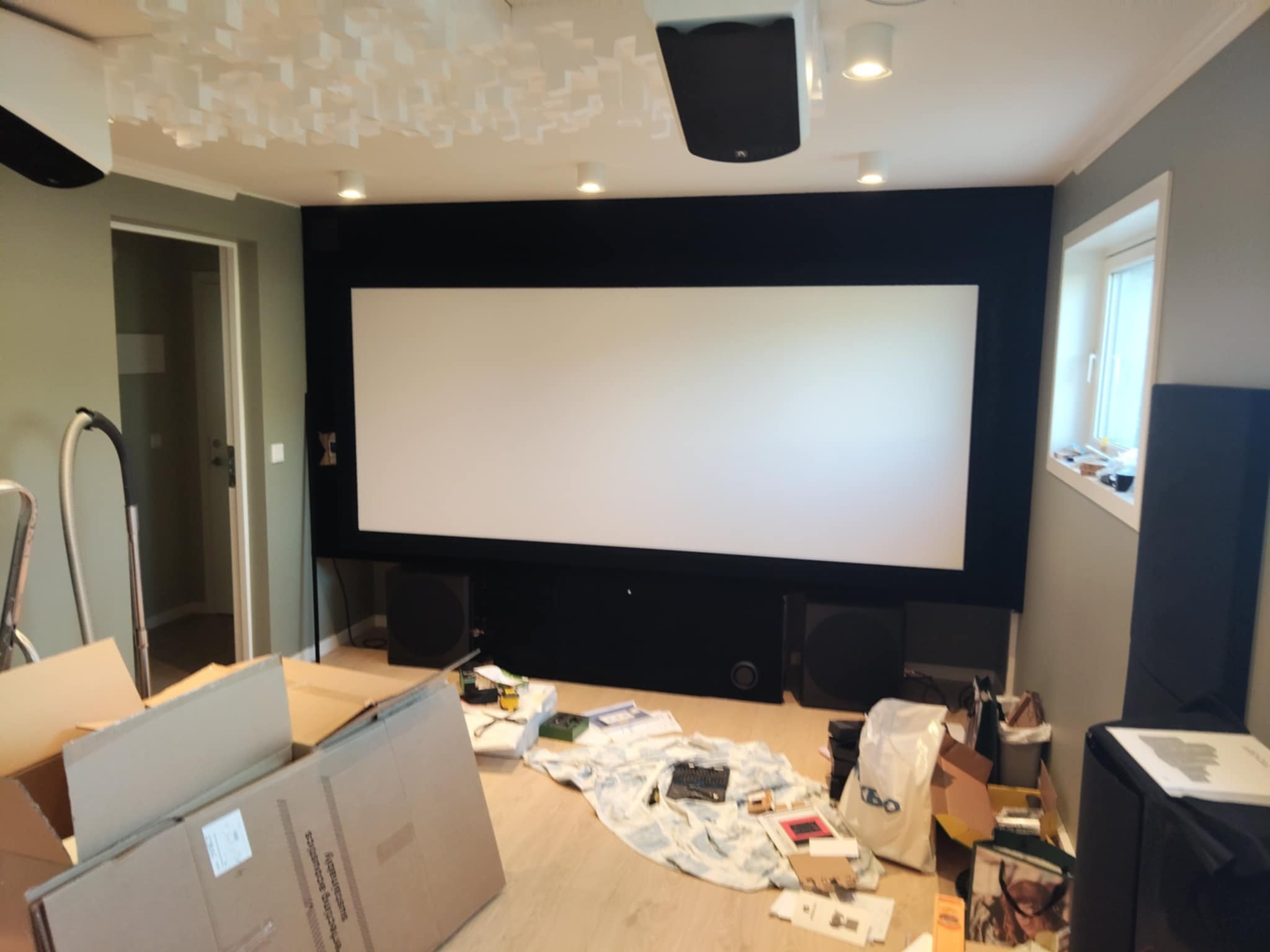 Wygłuszone pomieszczenie kina domowego z ekranem, głośnikami i panelami akustycznymi na suficie. Widoczne kartony i narzędzia sugerują etap wykończeniowy.