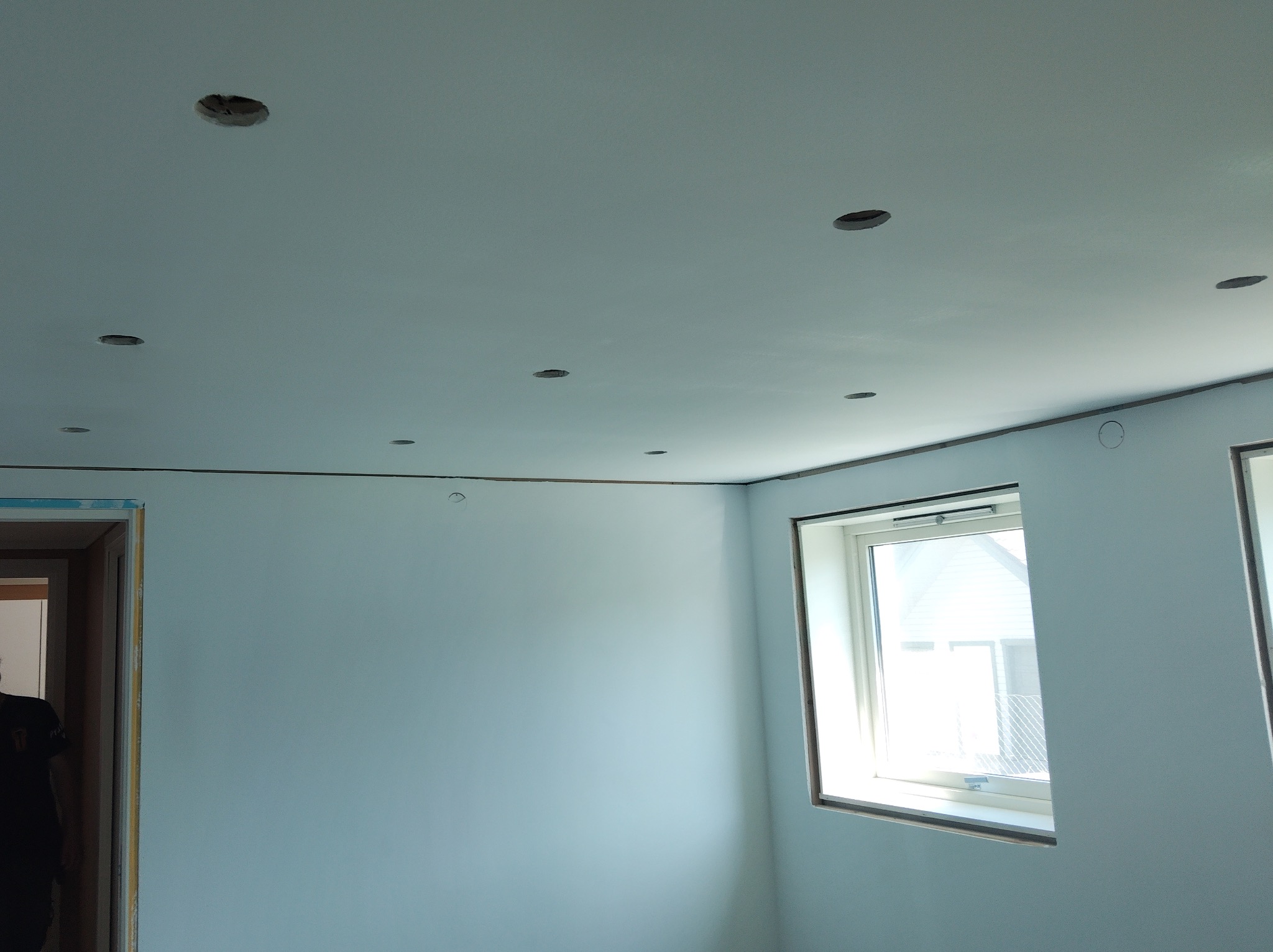 Jasne wnętrze po malowaniu ścian na delikatny odcień błękitu, z widocznymi otworami na oświetlenie sufitowe i oknem z widokiem na zewnątrz. Listwa przypodłogowa w kolorze brązowym.