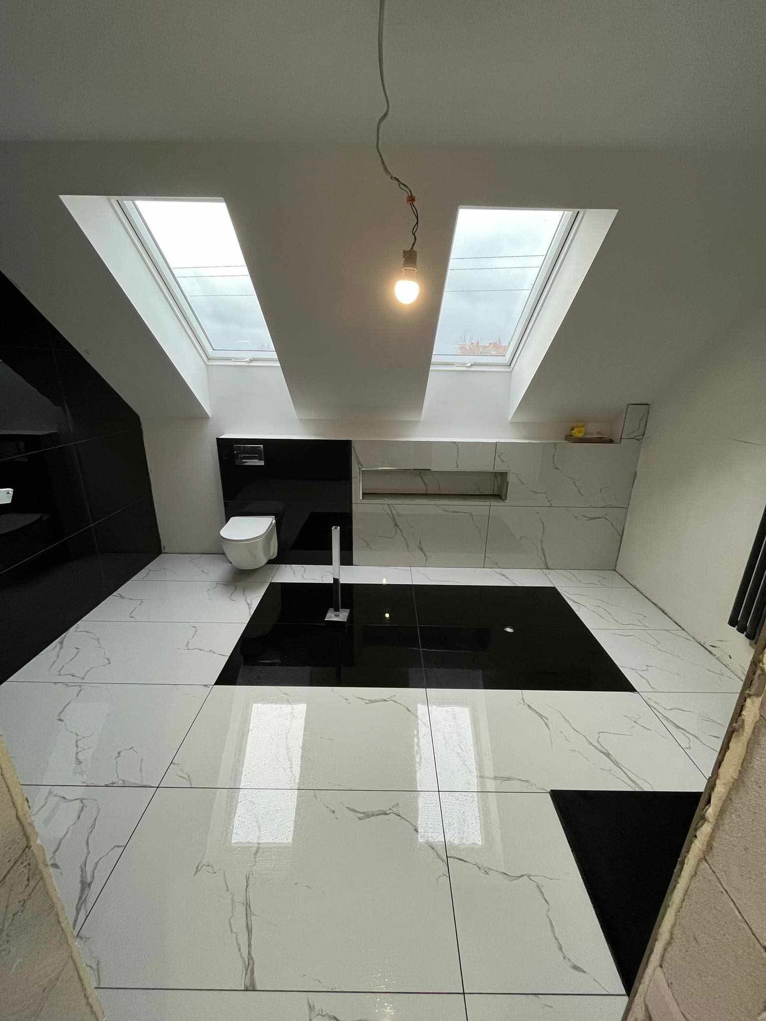 Nowoczesna łazienka na poddaszu z czarno-białymi płytkami imitującymi marmur, wiszącą toaletą i oknami dachowymi. Widoczny fragment niezakończonej podłogi.