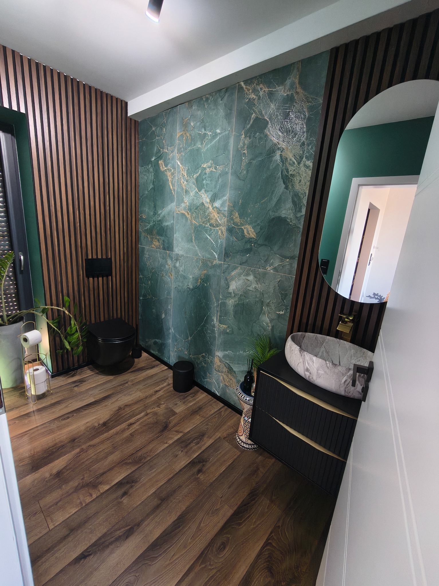 Nowoczesna łazienka z zielonymi płytkami imitującymi marmur, drewnianymi lamelami i umywalką z kamienia. Uwagę zwraca owalne lustro i czarna armatura.
