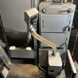 SZEWSKI-INSTAL - Szary piec Defro z podajnikiem i rurą spiro, widoczne elementy sterujące i otwarte drzwiczki. Ujęcie z bliska, w pomieszczeniu o surowym wykończeniu.