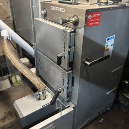 SZEWSKI-INSTAL - Szary, nowoczesny piec Defro z panelem sterowania i łańcuchem, widoczny w kotłowni. Na obudowie naklejka z numerem infolinii serwisowej.