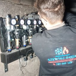 SZEWSKI-INSTAL - Instalator w trakcie montażu rozdzielacza ogrzewania podłogowego z manometrami, widoczny fragment ściany i logo firmy na bluzie montera.