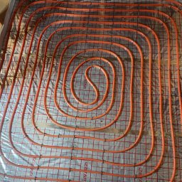 SZEWSKI-INSTAL - Pomarańczowe rury ogrzewania podłogowego ułożone spiralnie na folii izolacyjnej z siatką, przymocowane plastikowymi uchwytami. Widoczne drewniane elementy konstrukcyjne.