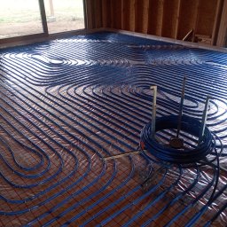 SZEWSKI-INSTAL - Niebieskie rury ogrzewania podłogowego ułożone na folii z siatką, widoczne narzędzia montażowe i fragment ściany z desek. Instalacja w trakcie realizacji.