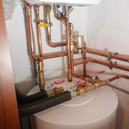 SZEWSKI-INSTAL - Instalacja hydrauliczna z miedzianymi rurami i zaworami Calido, zamontowana na białym zbiorniku w pomieszczeniu z białą, nierówną ścianą. Widoczne złącza i elementy mocujące.