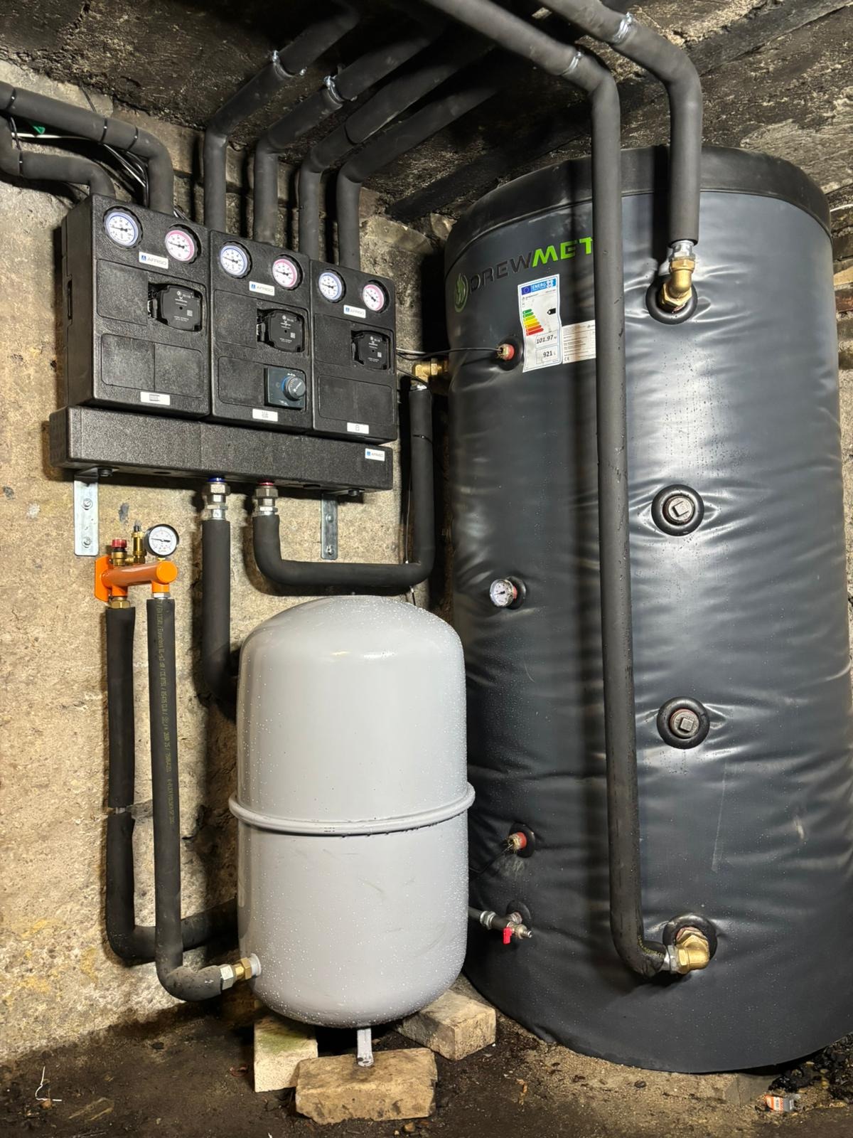 Zestaw instalacji grzewczej z czarnymi rurami, zbiornikiem akumulacyjnym i szarym naczyniem przeponowym, zamontowany w piwnicy. Widoczne manometry i zawory.