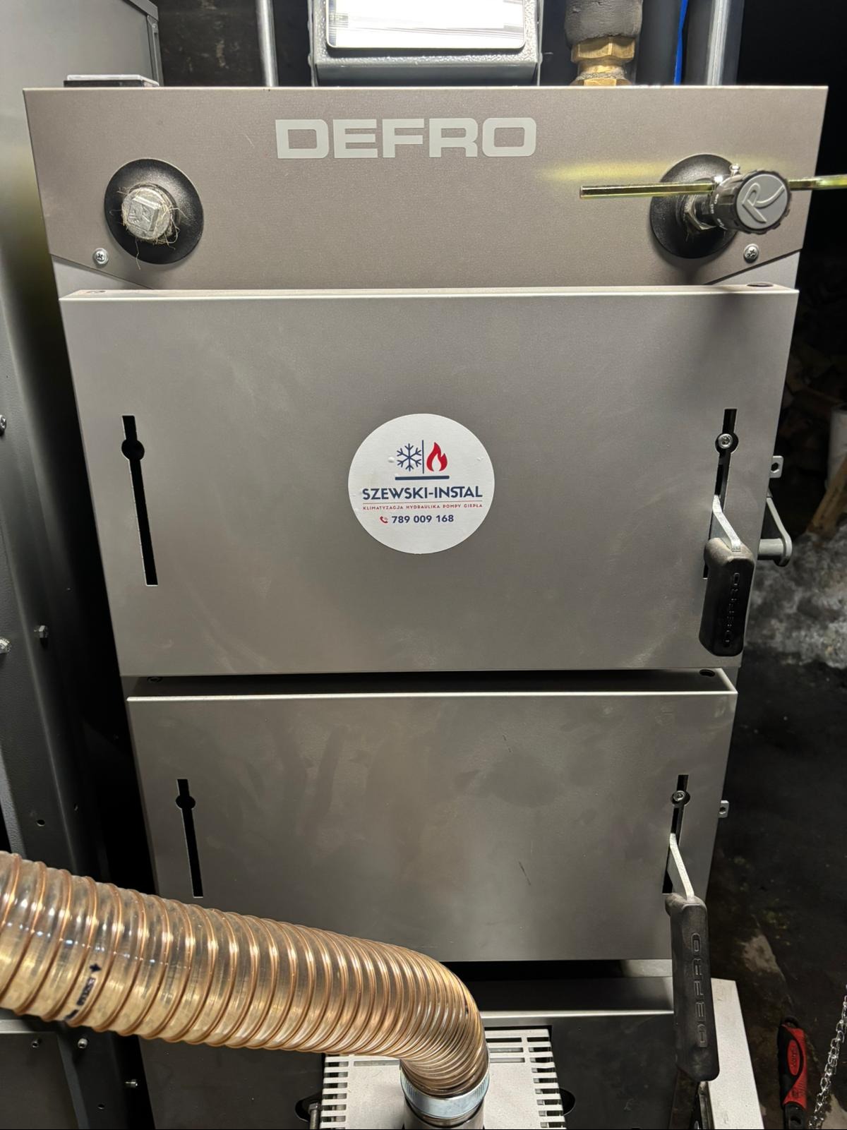 Szary piec DEFRO z naklejką firmy Szewski-Instal, widoczny fragment rury. Detal instalacji grzewczej, prezentacja kotła z bliska, z elementami regulacji i przyłączami.