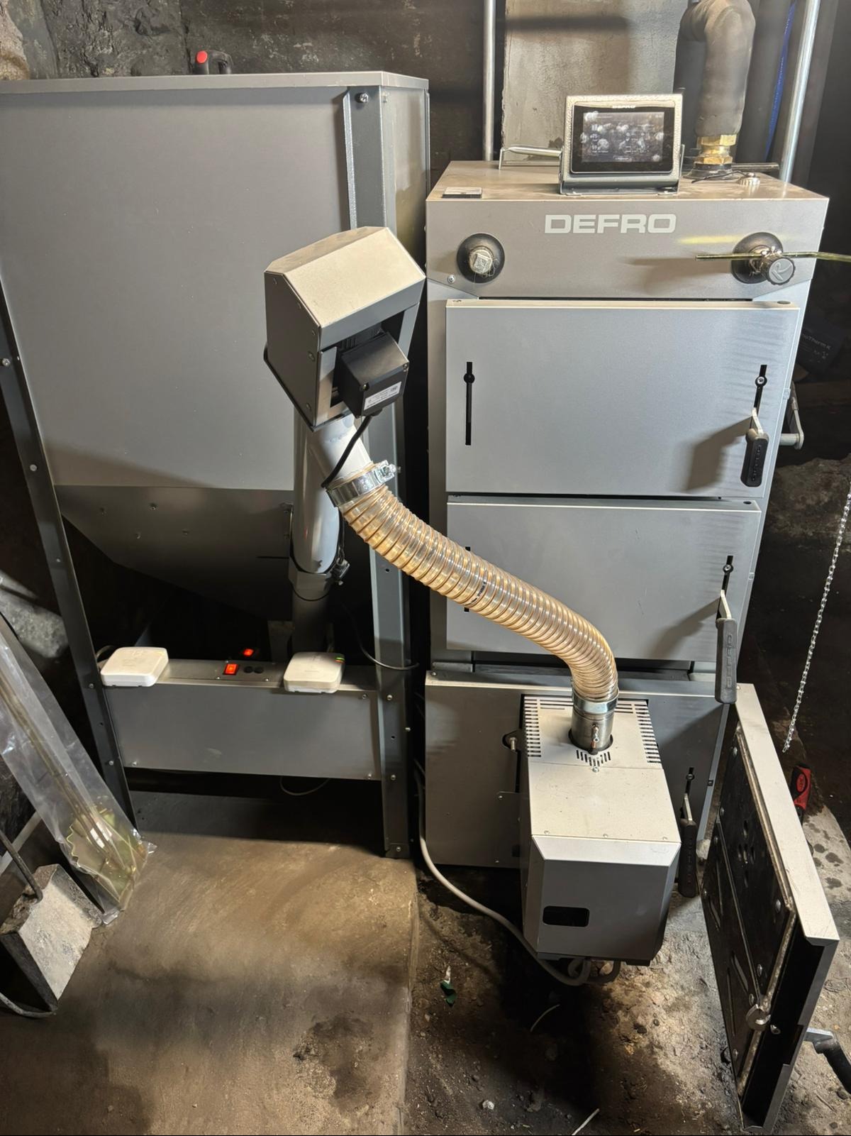 Szary piec Defro z podajnikiem i rurą spiro, widoczne elementy sterujące i otwarte drzwiczki. Ujęcie z bliska, w pomieszczeniu o surowym wykończeniu.